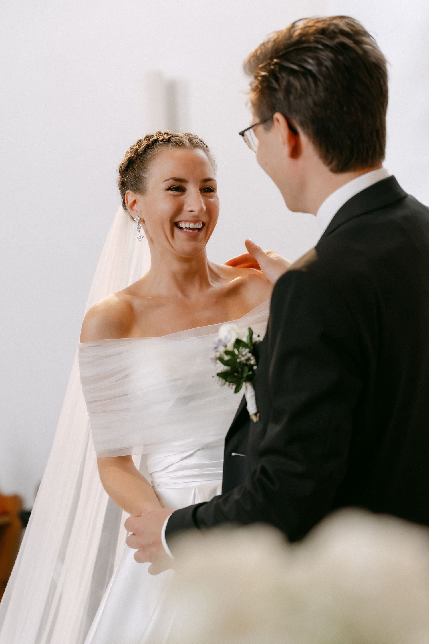 Smiling bride in an off-shoulder white wedding gown and veil laughs joyfully while facing a groom in a dark suit with a floral boutonniere.
