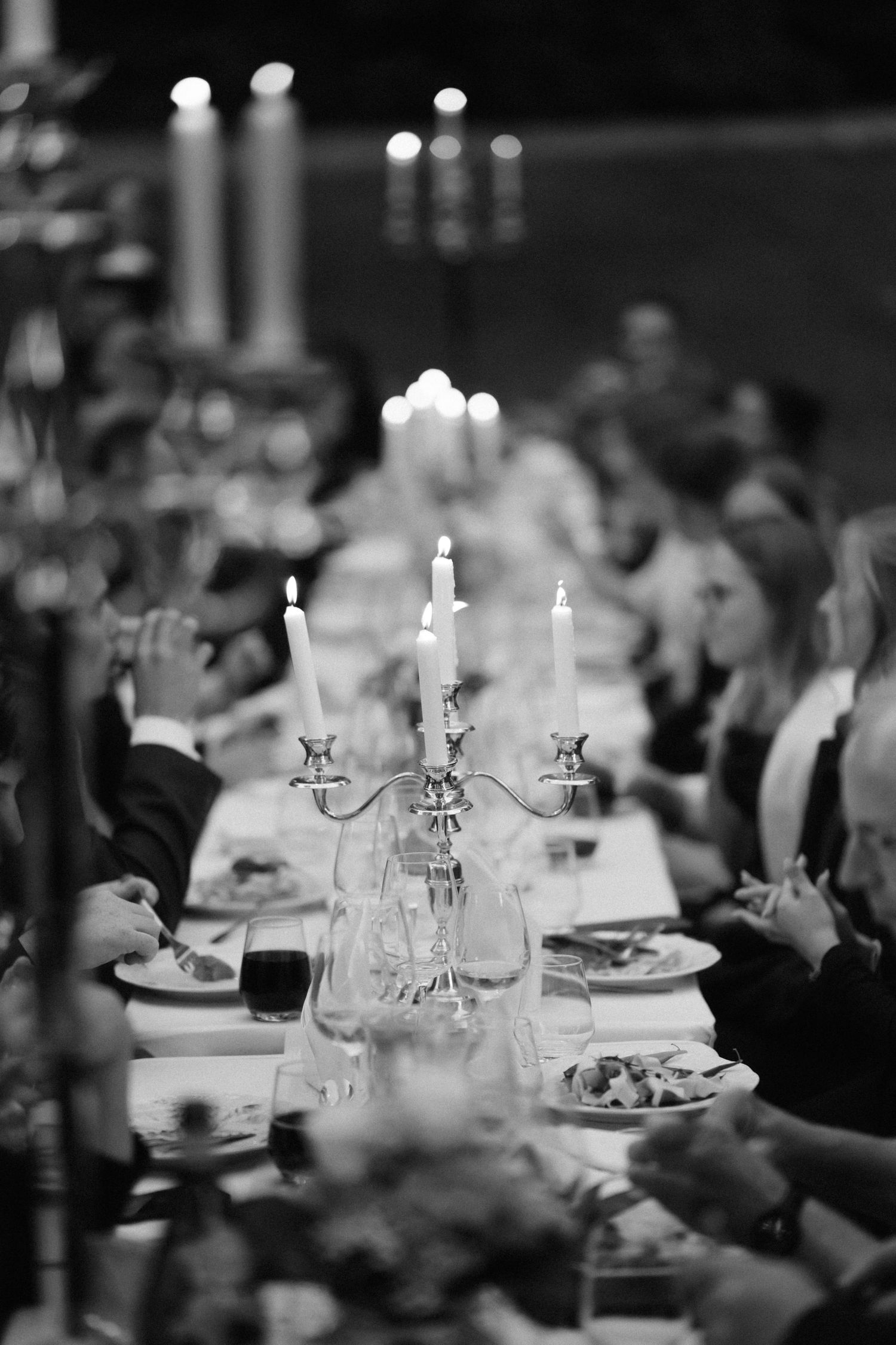 Black and white photo of guests dining at a long candlelit banquet table with silver candelabras, wine glasses, and plated dinner.
