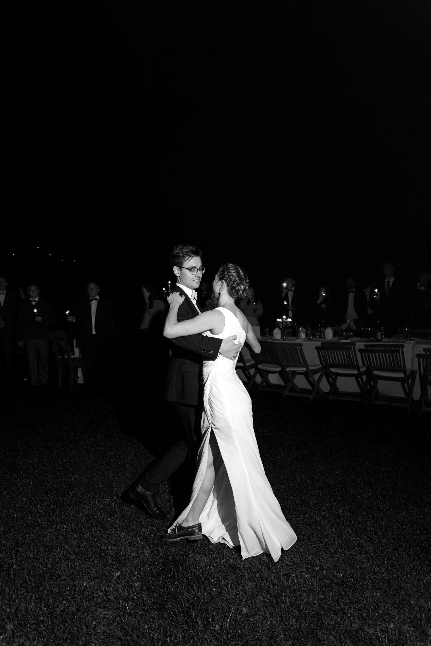 Black and white photo of a bride in a fitted gown and a groom in a suit sharing a first dance on a lawn at night as guests watch.
