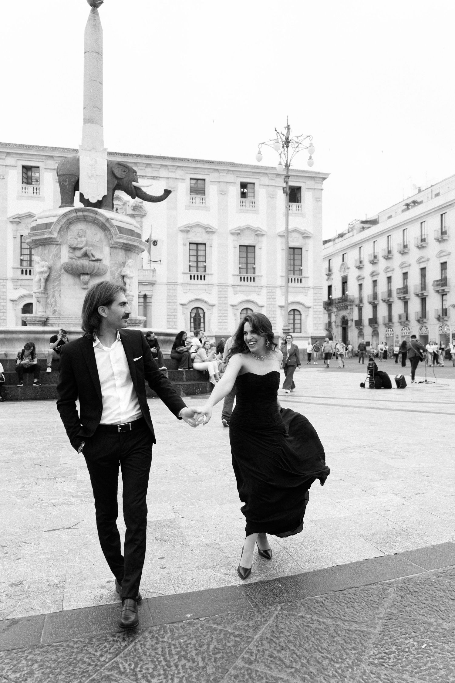 Elegant couple in formal attire holding hands while walking through a European plaza with a historic elephant fountain.
