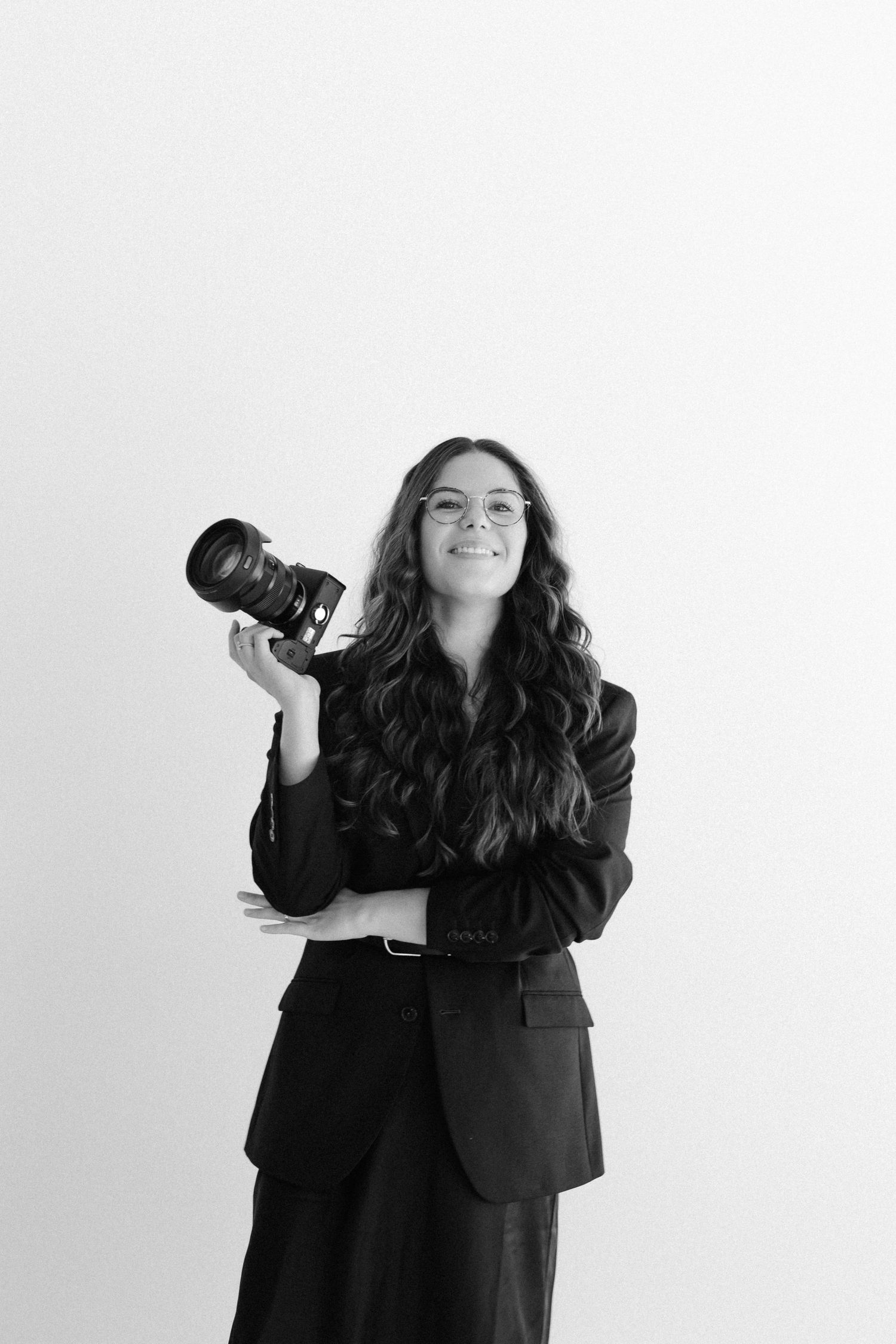 Black and white portrait of a young woman with curly hair, glasses, holding a professional camera, wearing a dark blazer.