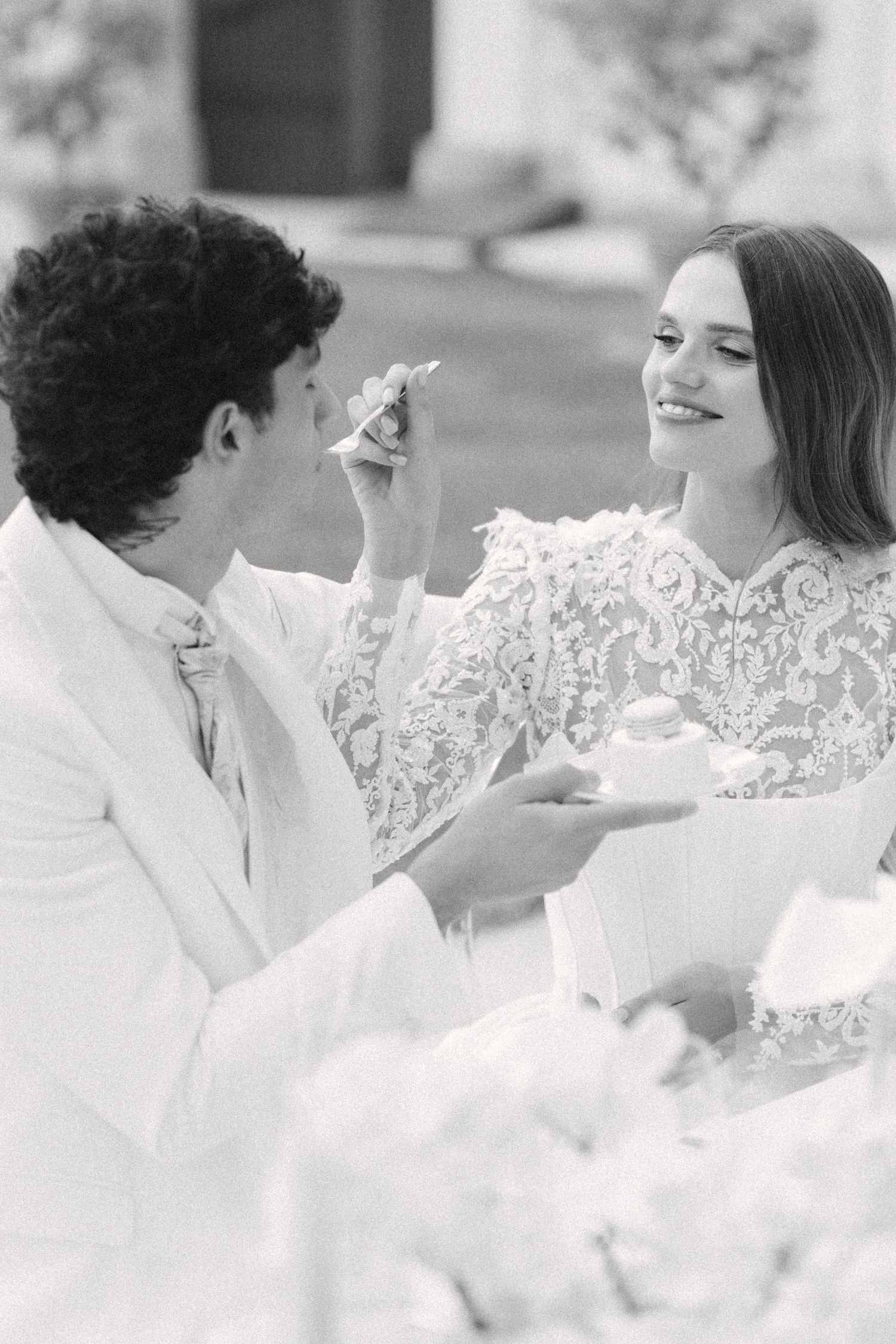 A black and white wedding photo showing a joyful moment as a couple in white attire share cake at their reception. Villa Mosconi Bertani