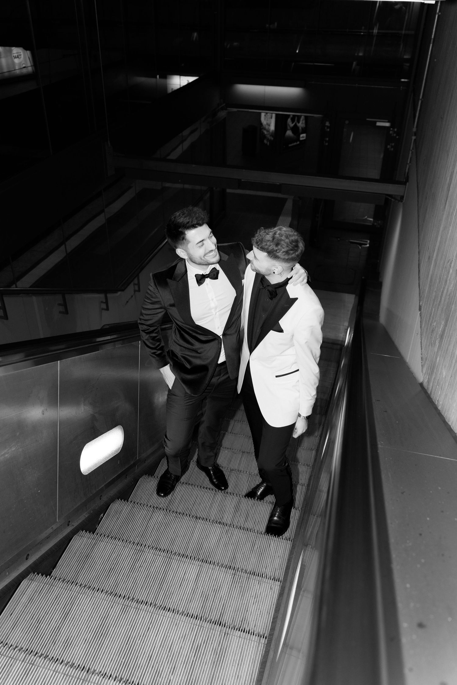 Two men in formal tuxedos and bow ties standing together on an escalator in a black and white photograph.