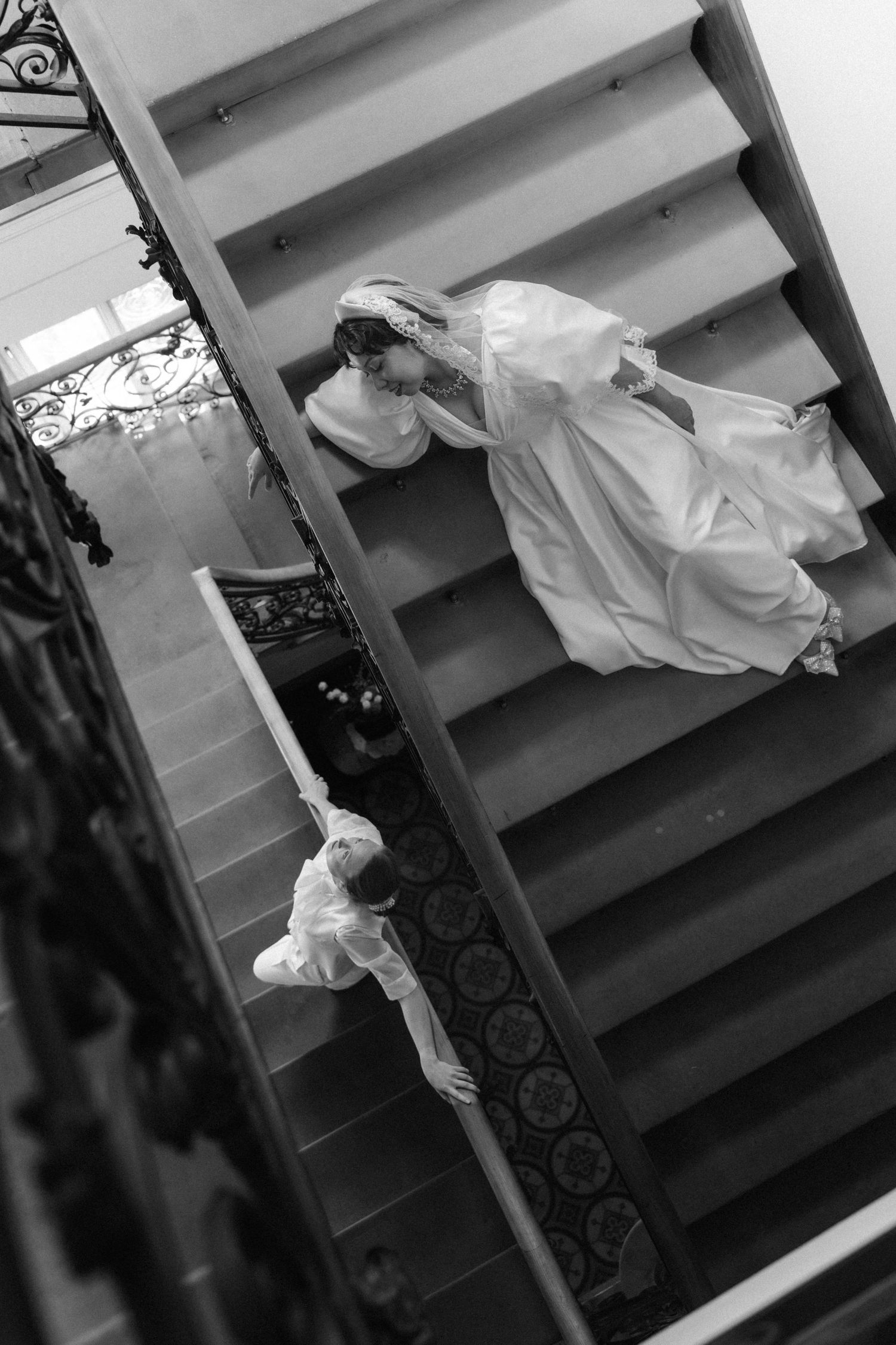 Black and white photo of a bride in a white gown and veil descending an elegant staircase with an attendant below.