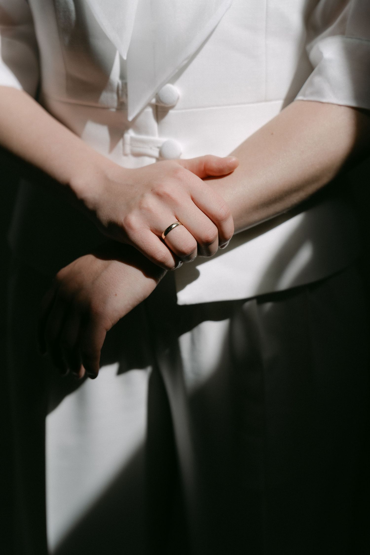Close-up of two hands clasped together, one wearing a gold ring, against a black skirt and white blouse.