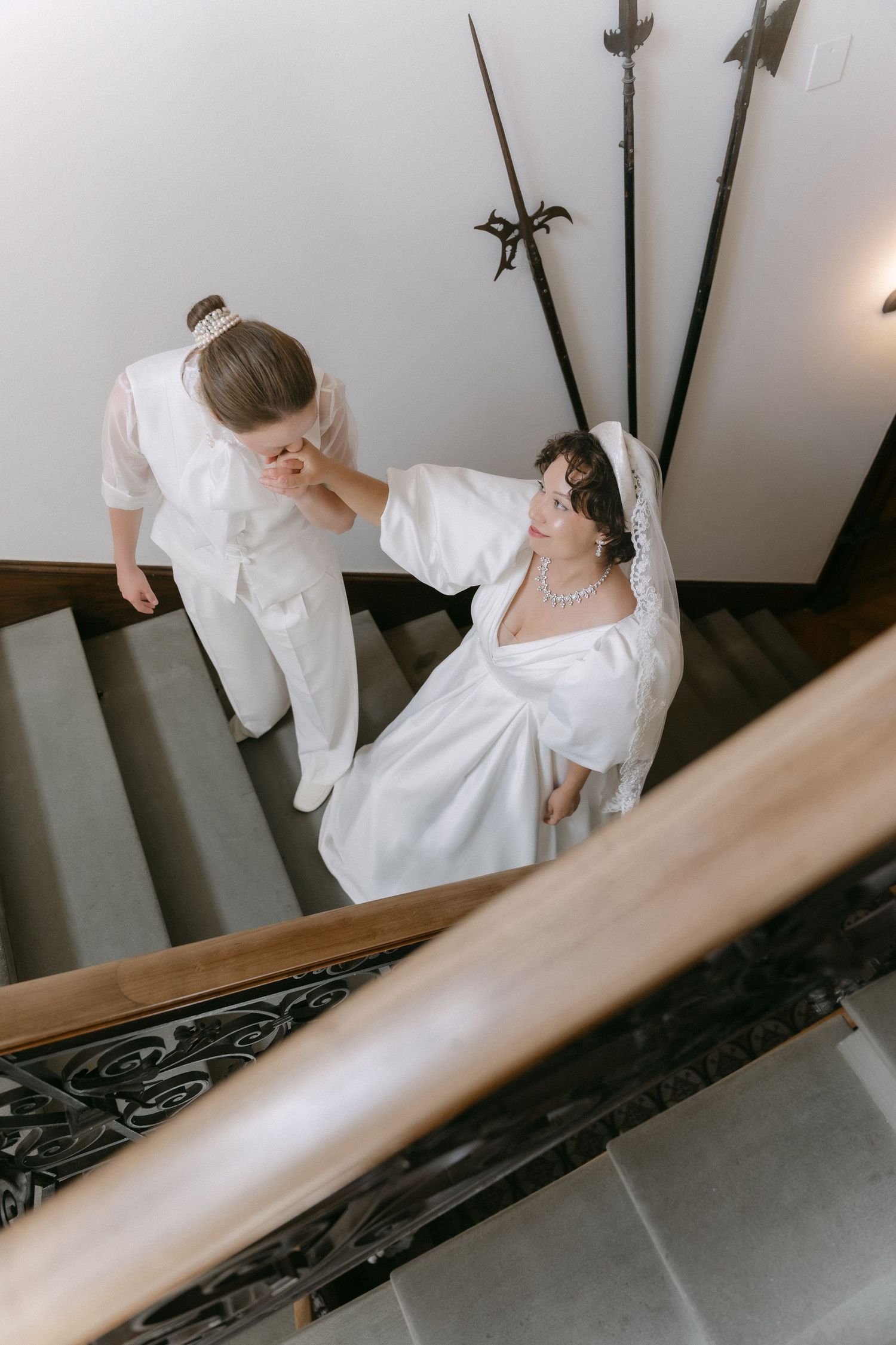 Groom kisses bride's hand on elegant staircase, both in white wedding attire with decorative swords in background.