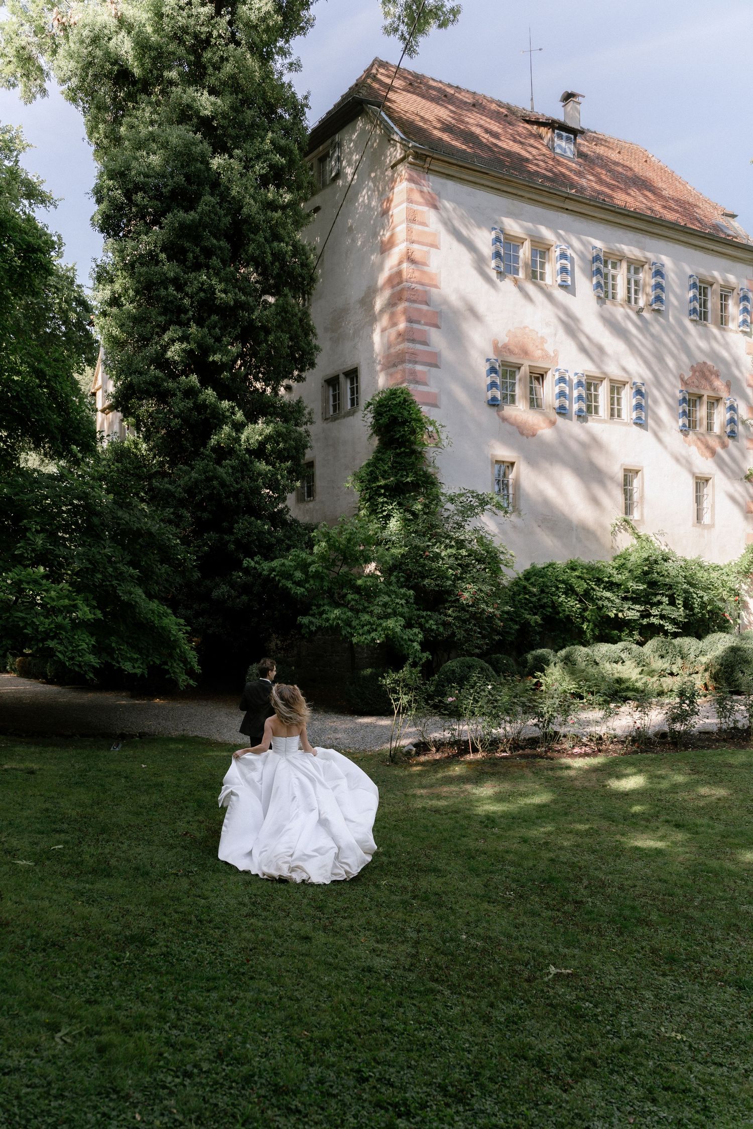 Bride in a flowing white ball gown walks across a sunlit lawn with her groom toward a historic castle with shuttered windows and red roof.