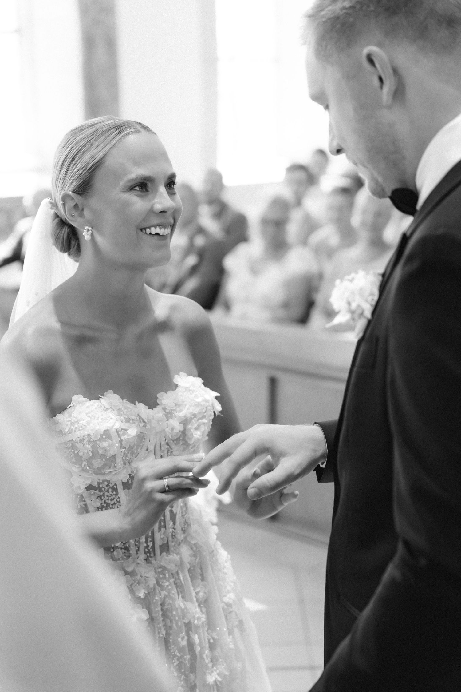 Smiling bride in a lace strapless gown exchanges wedding rings with the groom in a black tuxedo as guests watch during the ceremony.