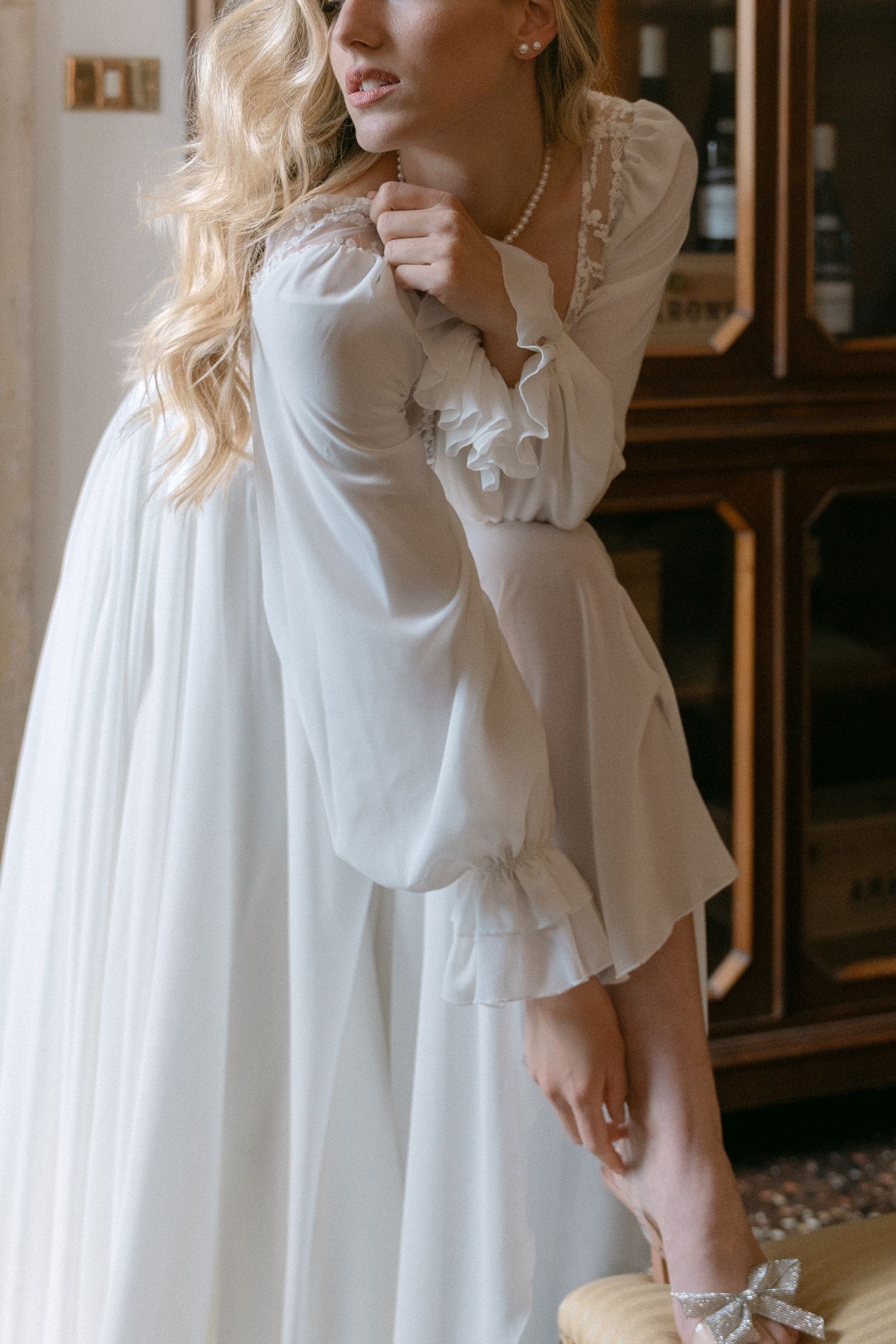 Woman in flowy white vintage-style dress with ruffled details poses gracefully in an elegant interior setting. Wedding Verona