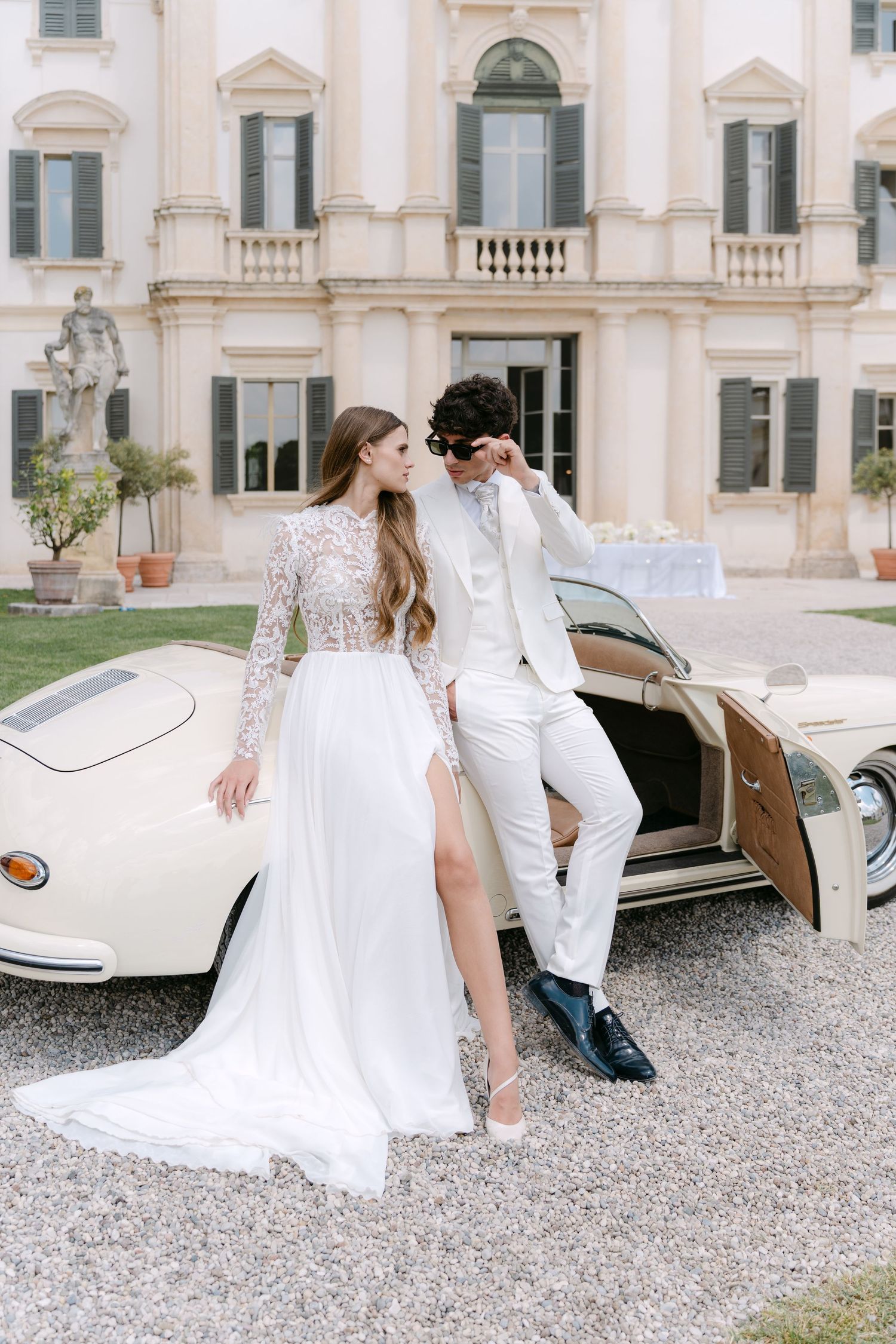 Elegant couple in white wedding attire pose beside vintage cream-colored car outside grand Italian villa. Villa Mosconi Bertani