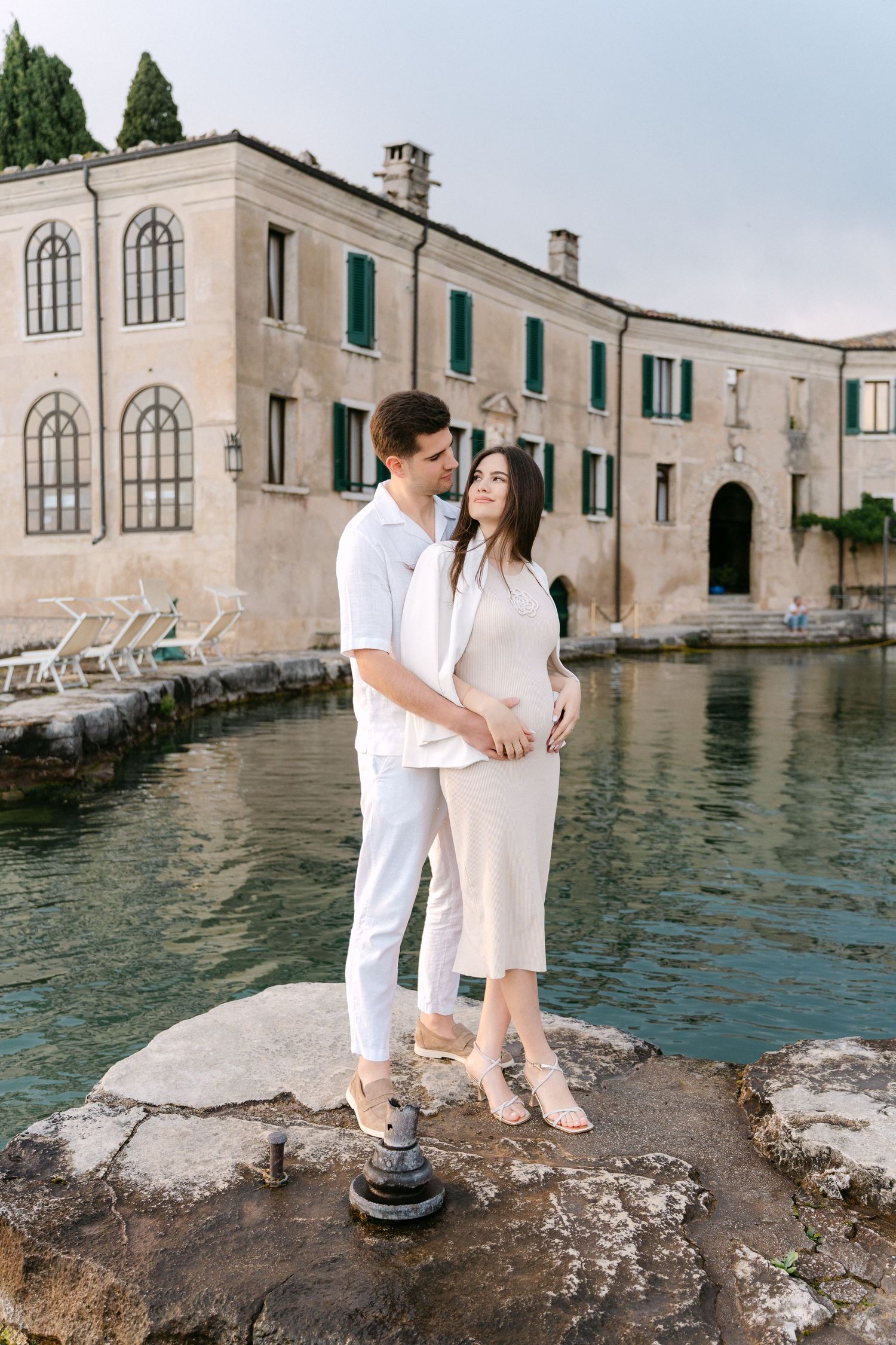 Couple in white outfits sitting by the water's edge at a historic Mediterranean villa with stone architecture.