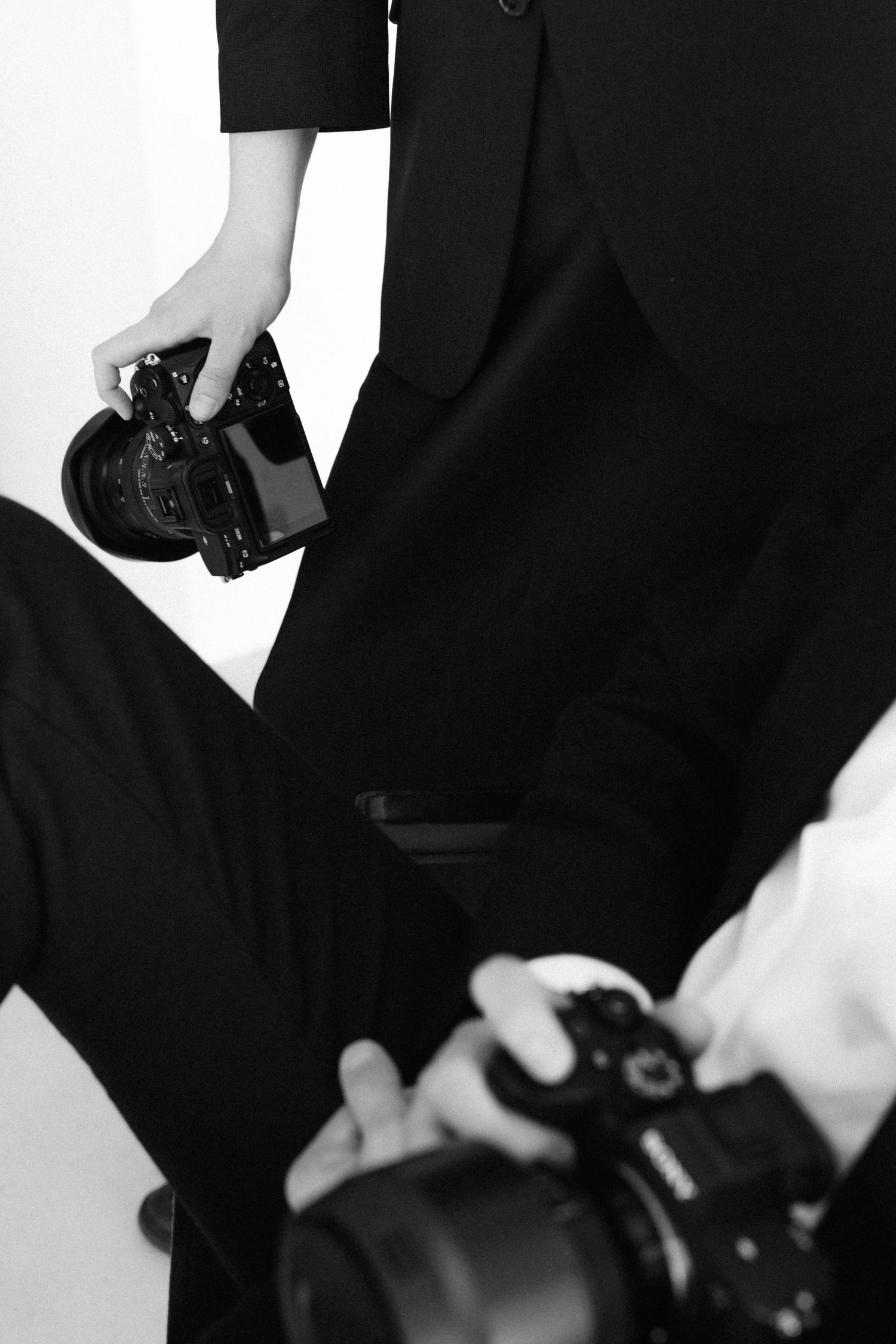 Black and white photo of two photographers in black suits holding professional DSLR cameras in a studio setting.
