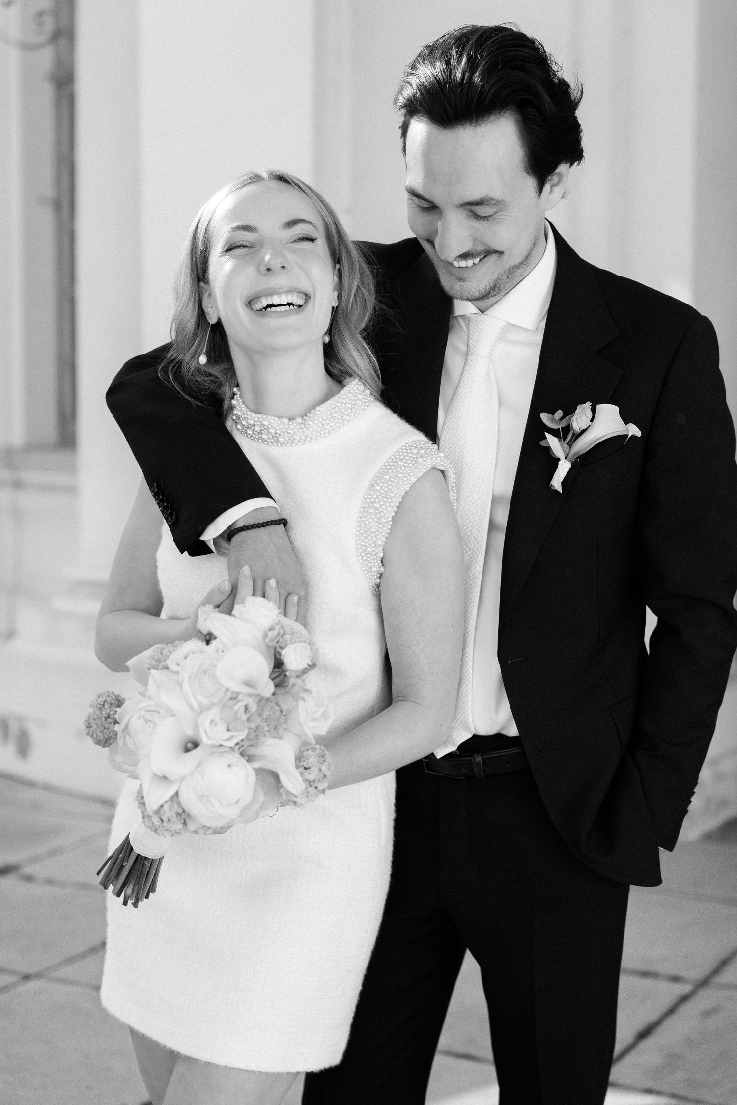 Black and white photo of a laughing bride in short dress holding bouquet, embraced by smiling groom in suit with boutonniere.