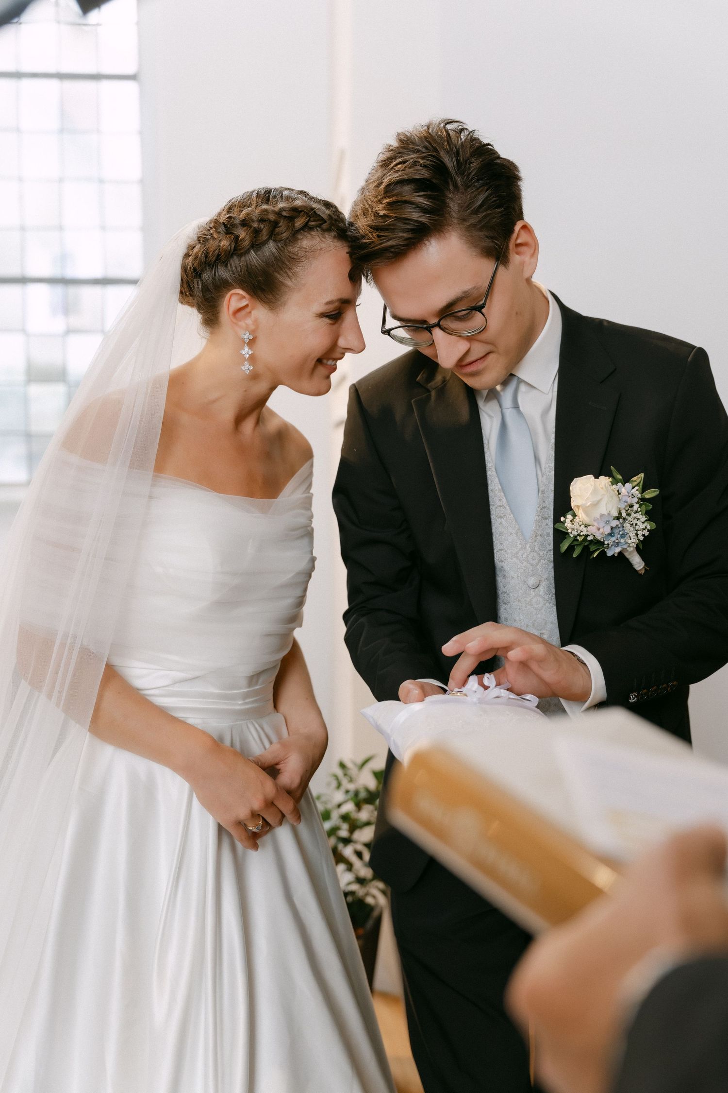Bride in a white satin gown and veil smiles beside a groom in glasses and a black suit as they exchange rings during a wedding ceremony.