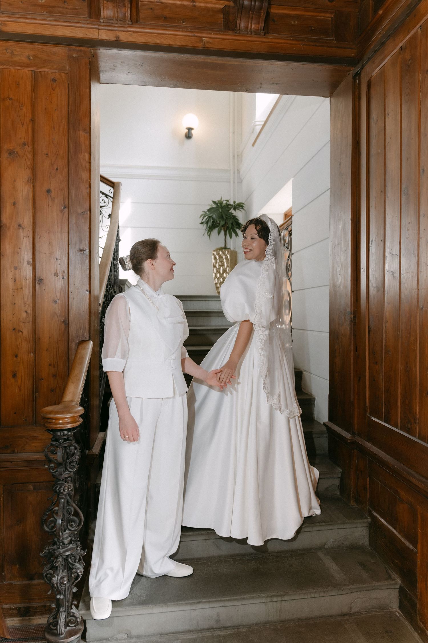 Two women in white outfits holding hands on elegant wooden staircase in a classic interior setting.