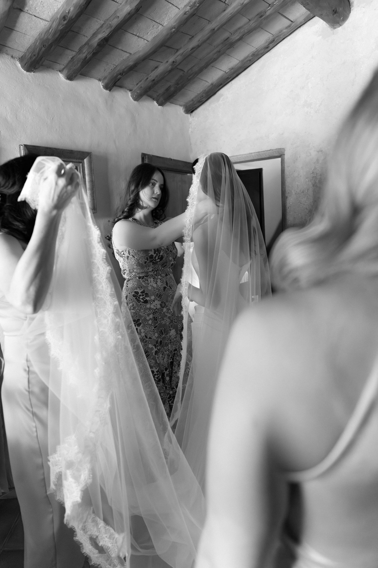 Bridesmaids helping bride with veil in rustic room, black and white wedding preparation photo.