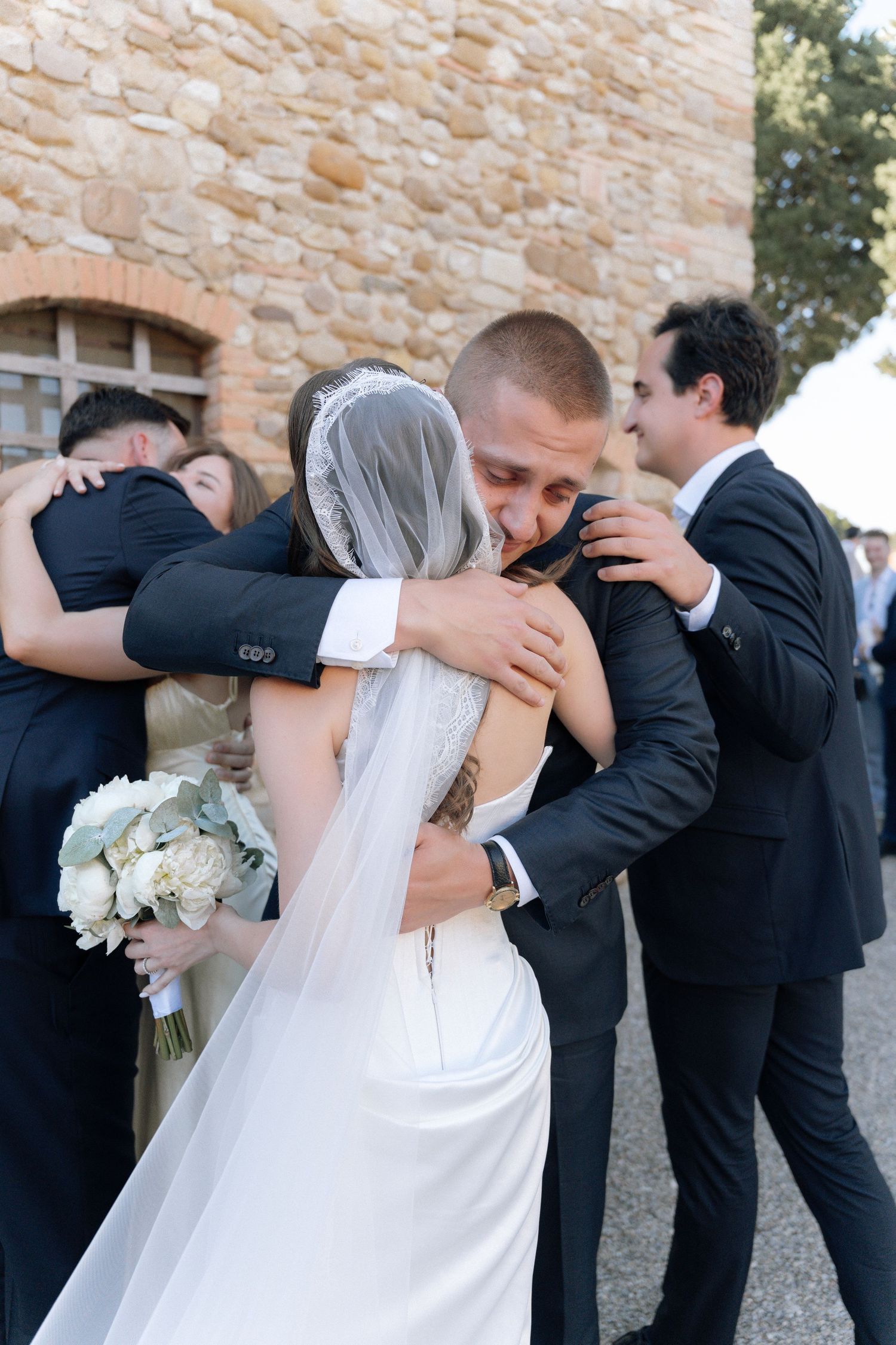 Bride and groom share an emotional hug with wedding guests outside a stone building after their ceremony.