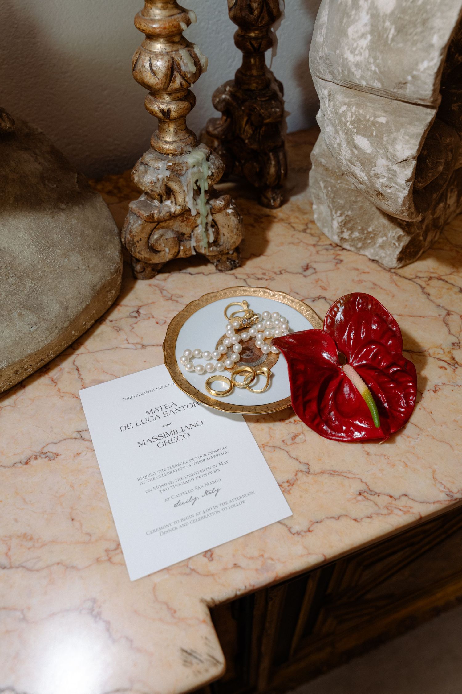 Marble table with wedding invitation, pearl necklace on gold dish, red anthurium flower, and ornate golden candlesticks.