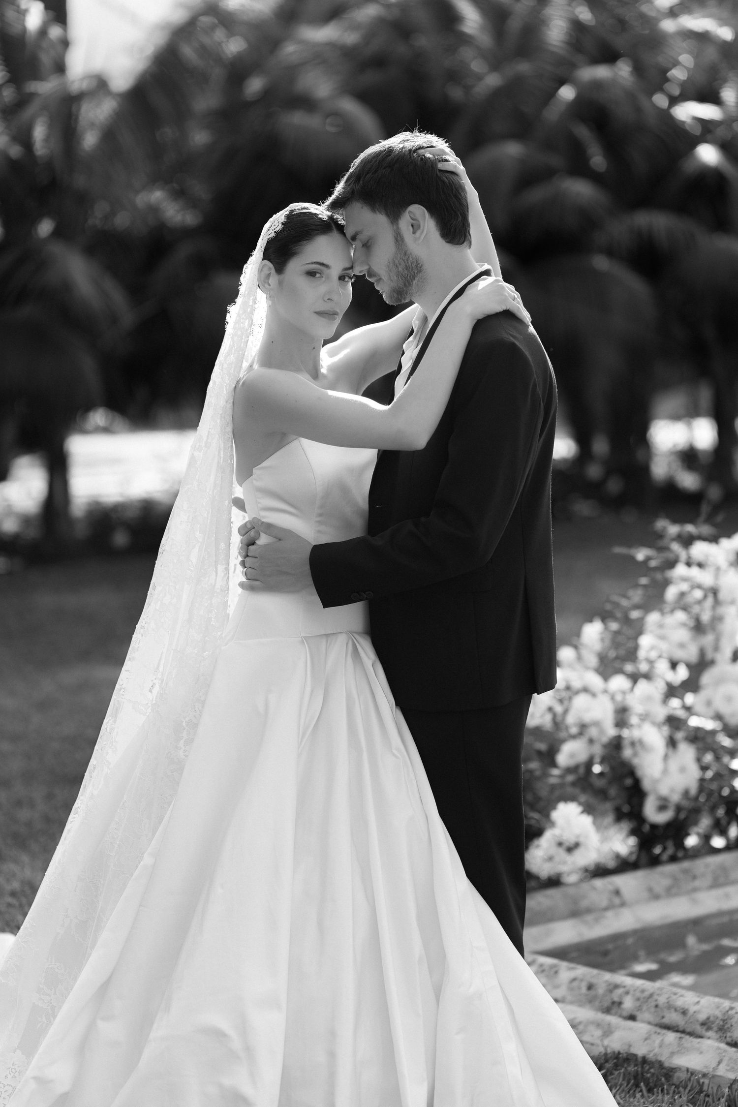 Black and white photo of a bride in white gown and groom in tuxedo embracing outdoors with palm trees.