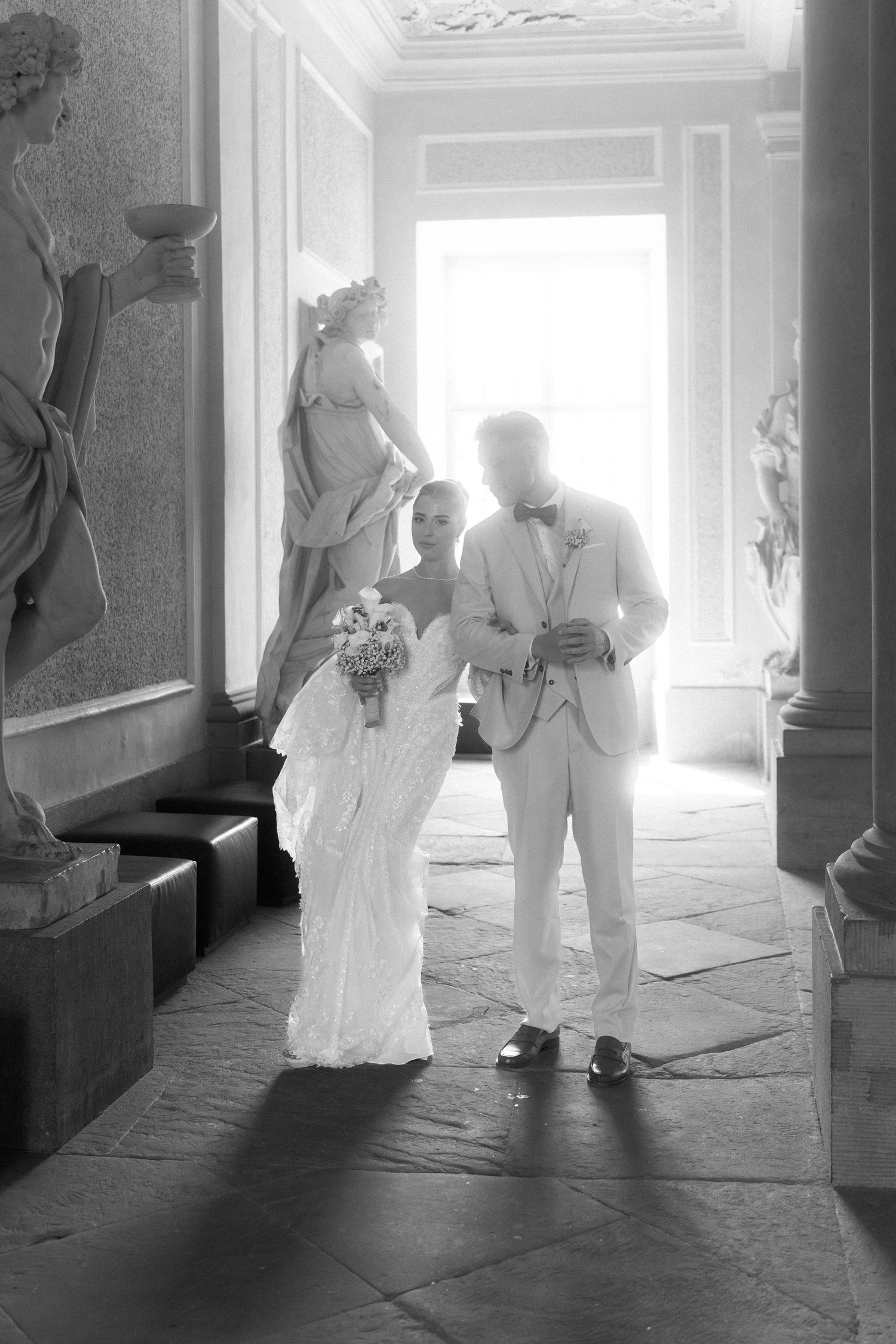 Black and white sequence of figures in white clothing standing in a sunlit hallway with stone columns and flooring. Schloss Ludwigsburg