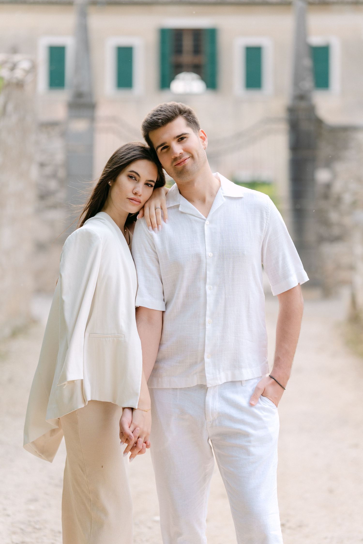 A romantic couple in white outfits poses together against a light stone wall backdrop in a series of intimate photos.