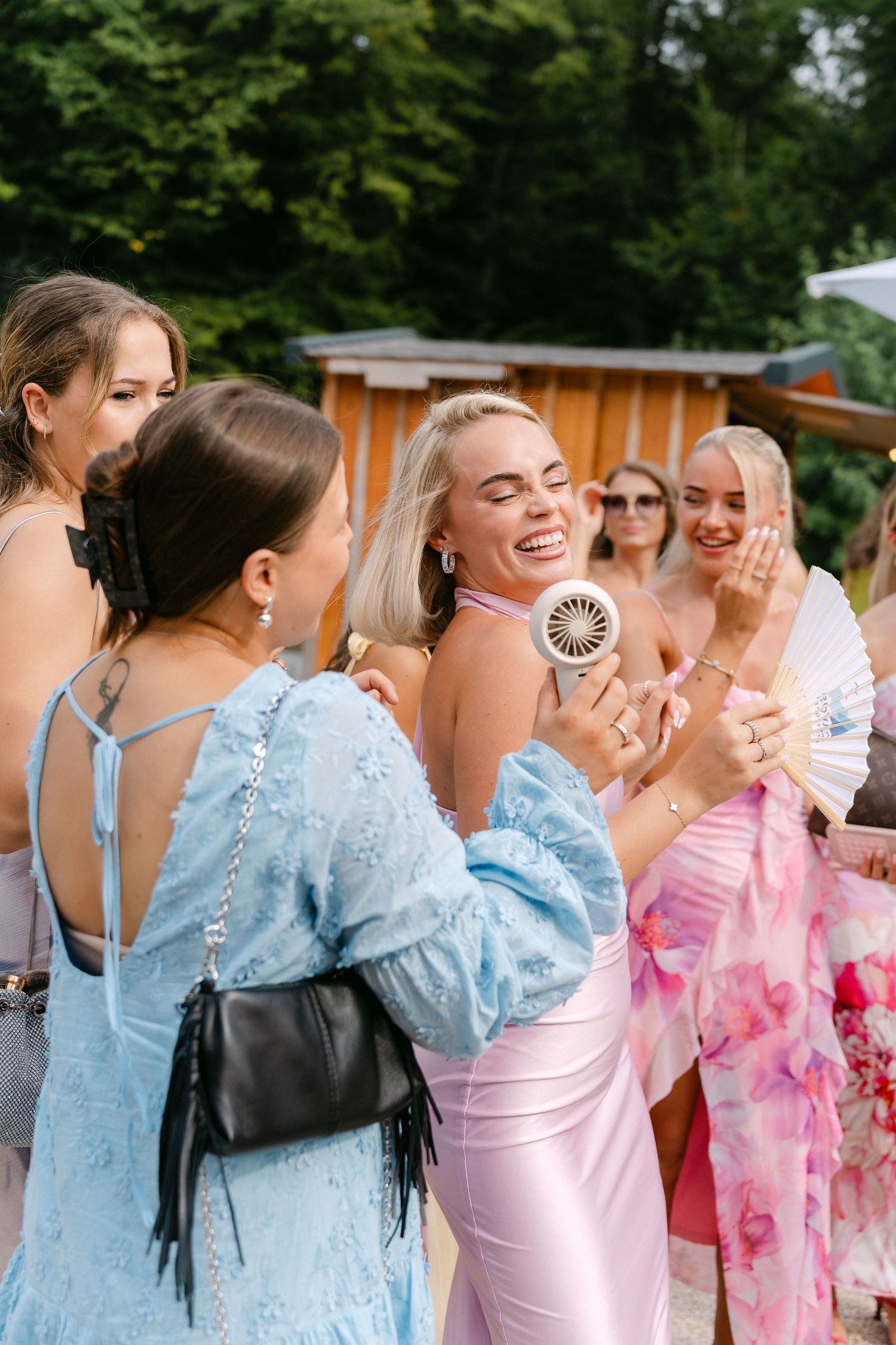 A group of wedding guests in blue and pink dresses share joyful moments and laughter during an outdoor celebration.