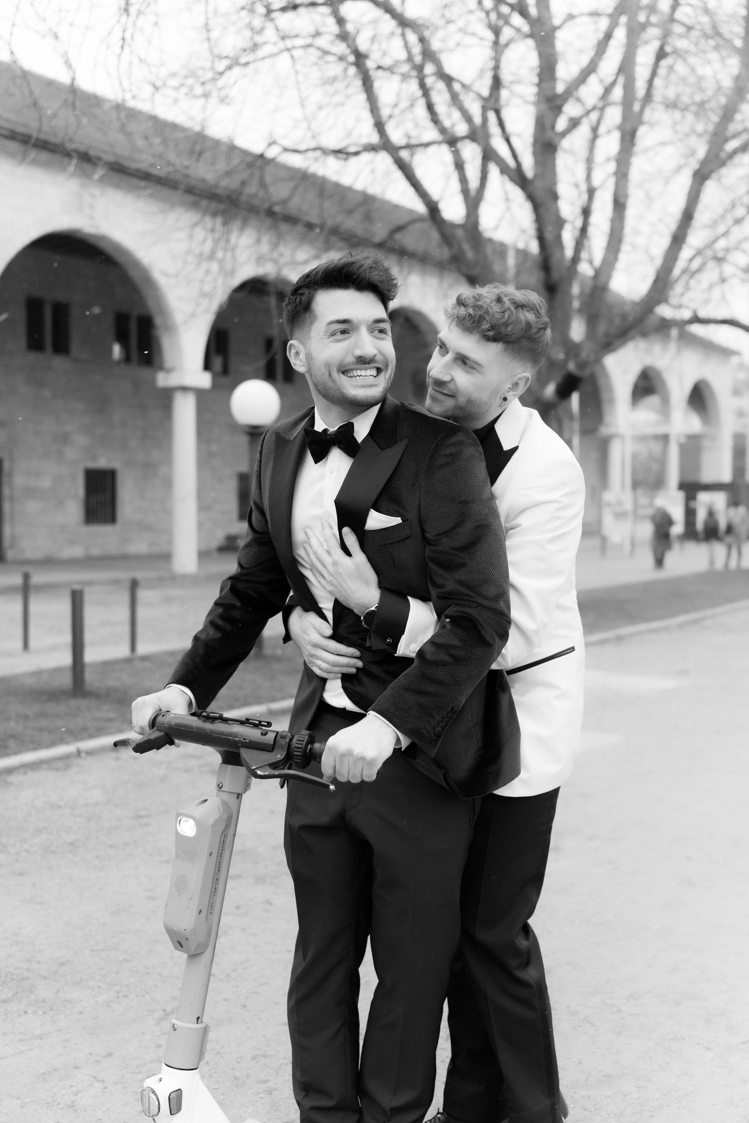 Two people in formal attire sharing a joyful moment on an electric scooter in front of an arched building.