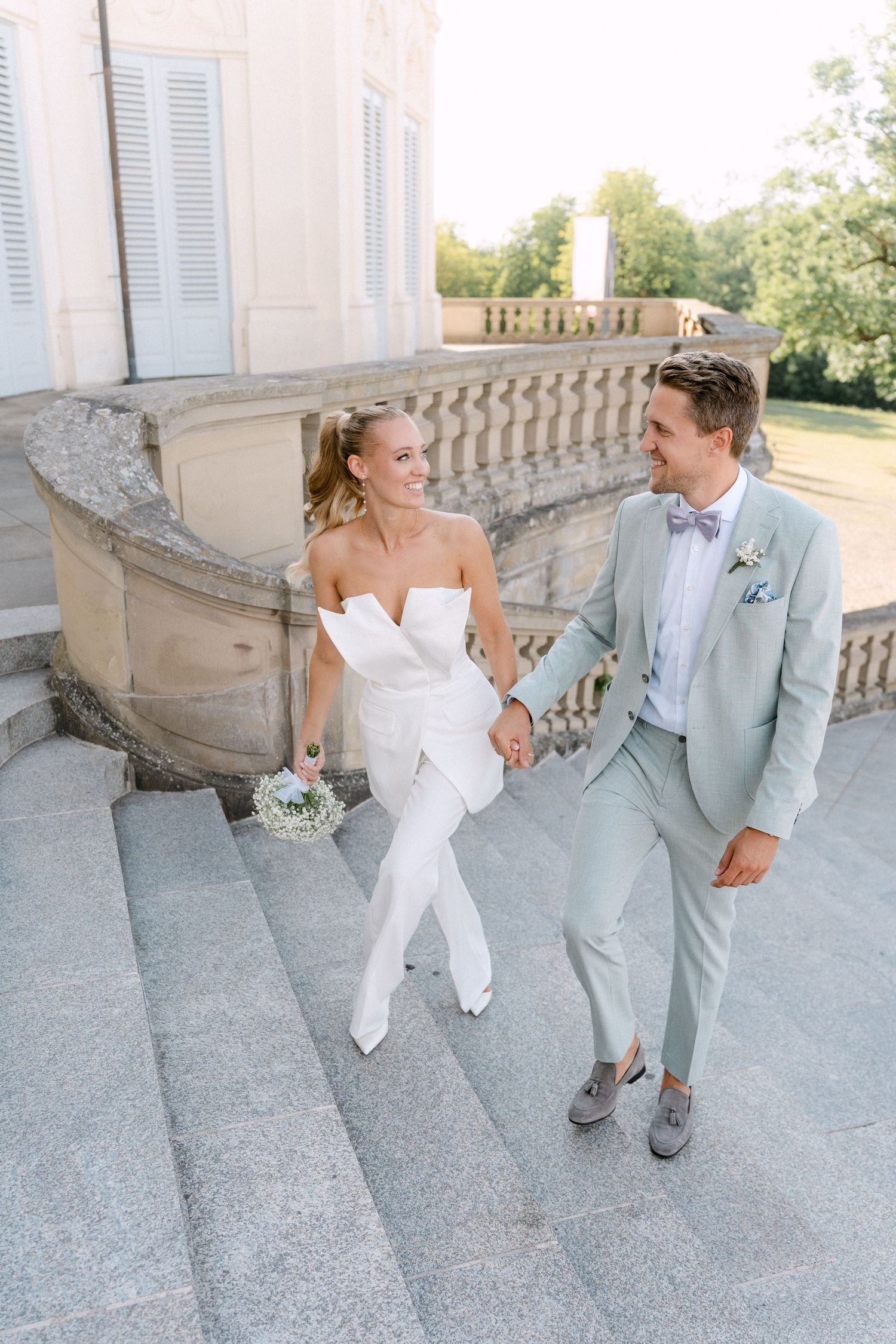A bride in a white jumpsuit and groom in light gray suit hold hands on an elegant stone terrace. Schloss Solitude
