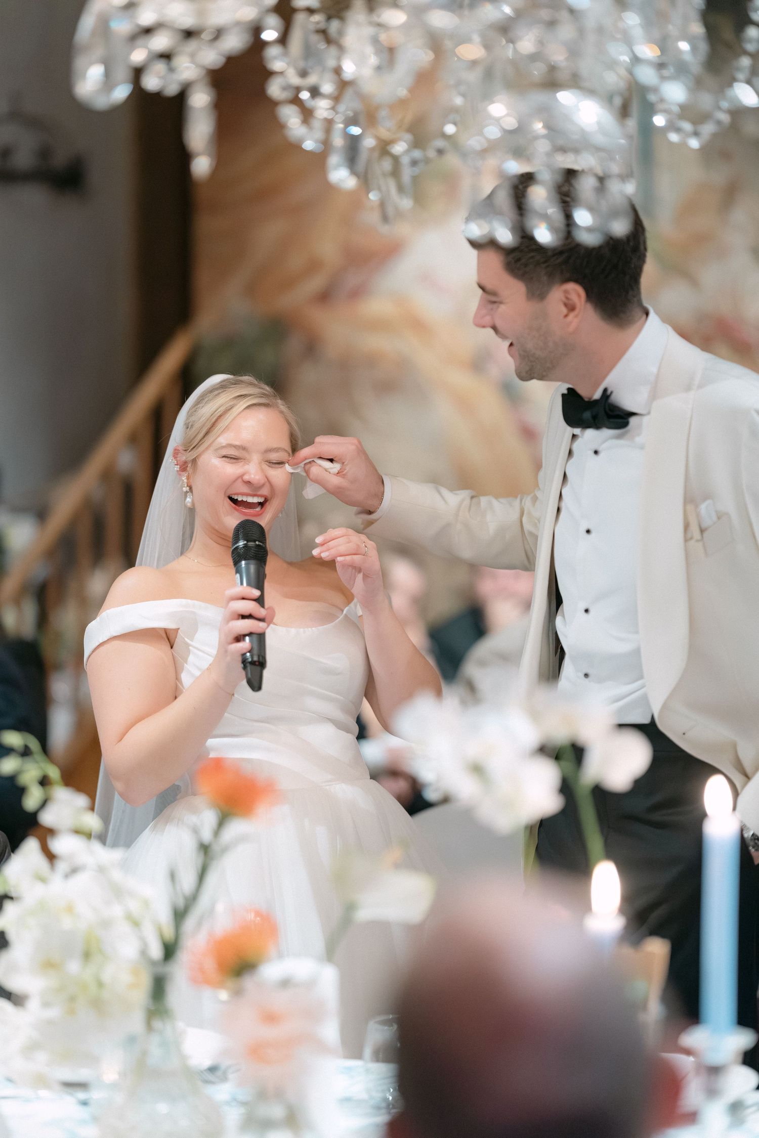 A joyful moment captured during a wedding reception as the couple shares laughter under elegant crystal chandeliers.