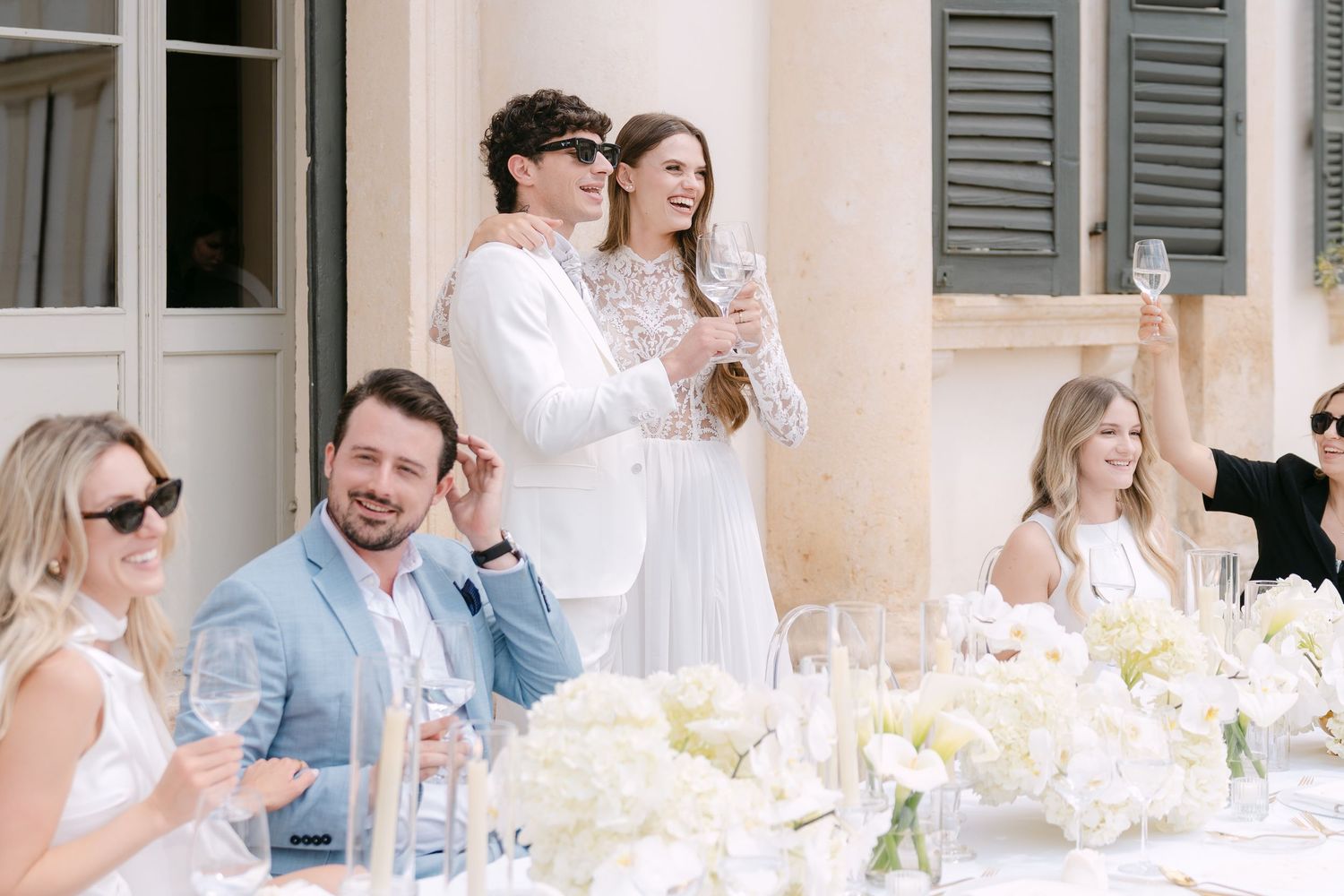 Guests enjoy drinks at an elegant outdoor wedding reception with white floral centerpieces and table settings. Villa Mosconi Bertani