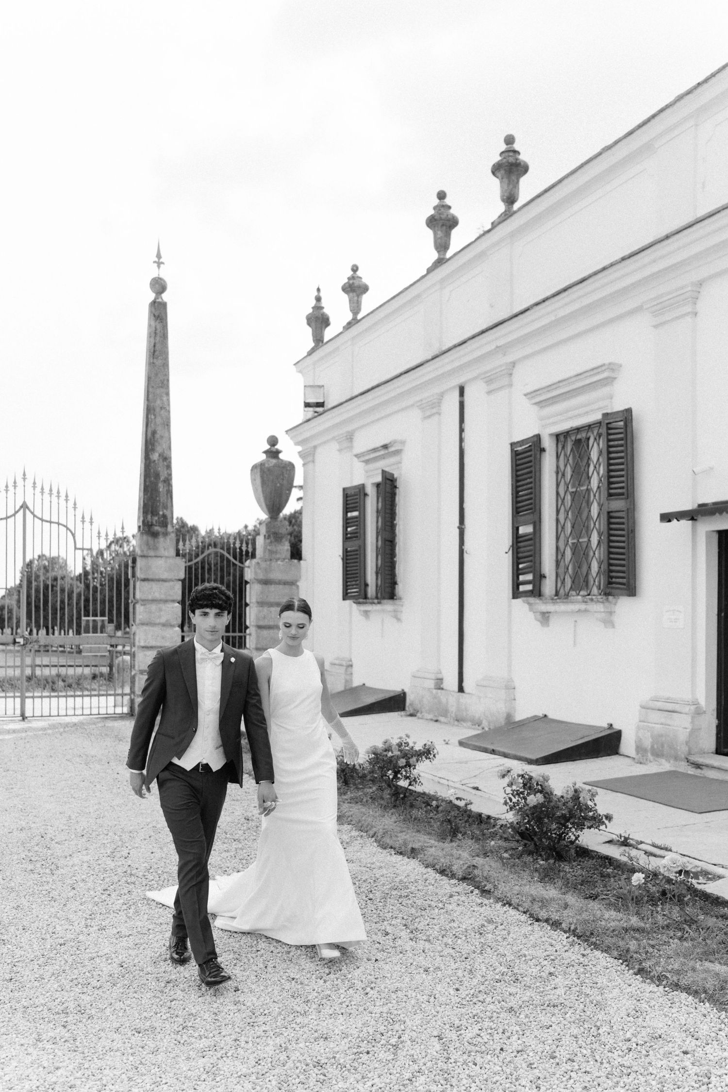 A couple walks hand in hand along a gravel path beside a white Mediterranean-style building with black shutters. Villa Mosconi Bertani