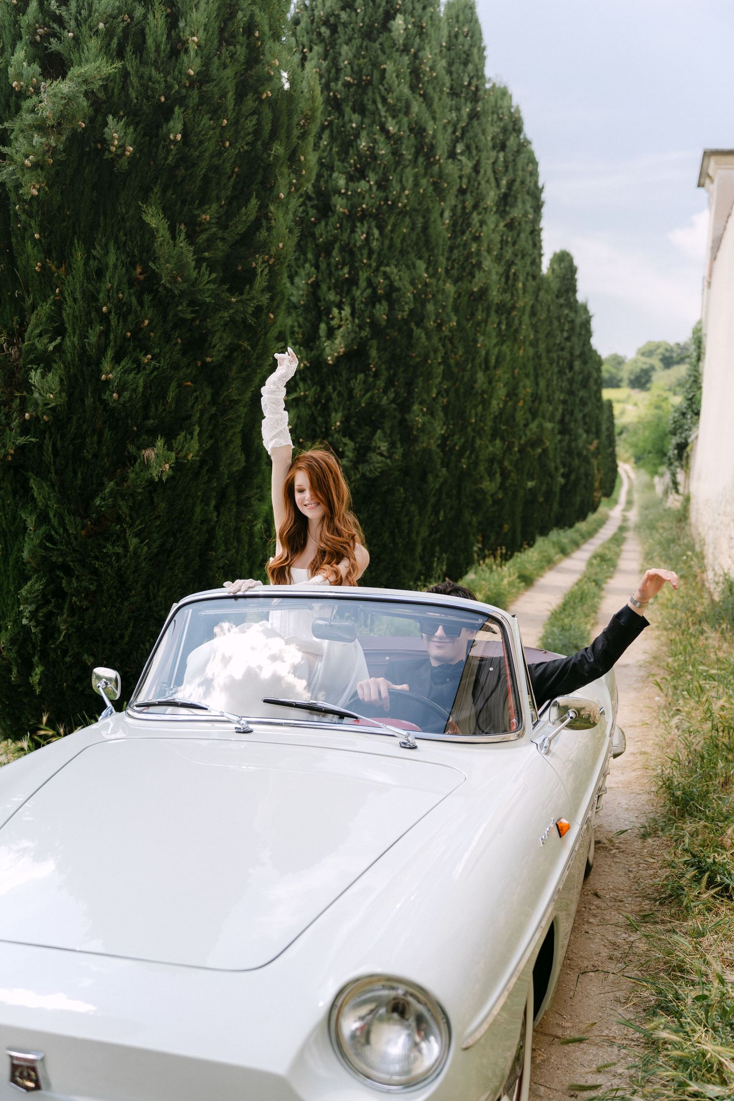 A white vintage convertible car drives along a tree-lined road in the Italian countryside.