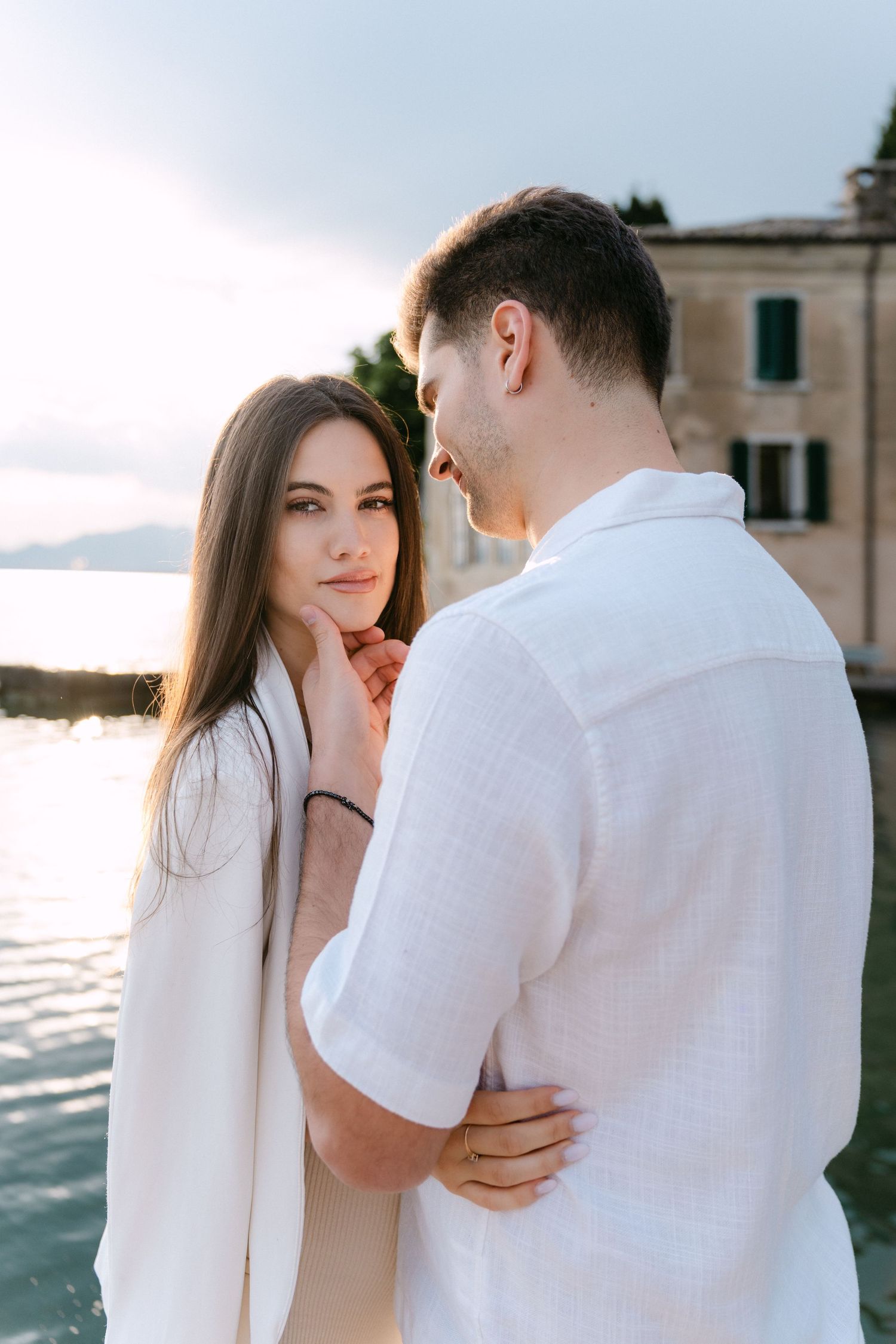 A romantic couple in white clothing embraces by the water during a seaside photoshoot at sunset.