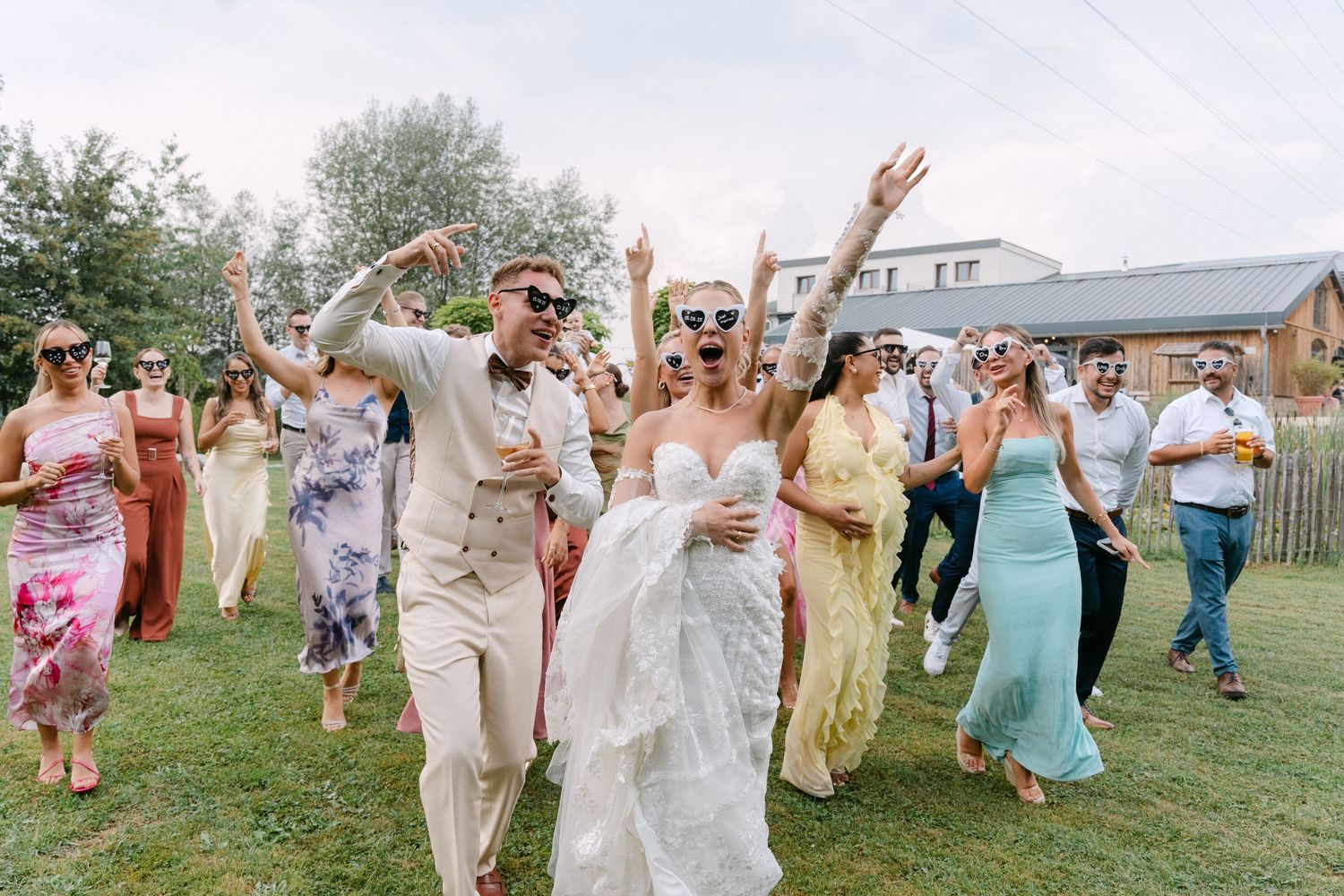 A group of wedding guests dance together outdoors on a grassy lawn during a summer celebration.
