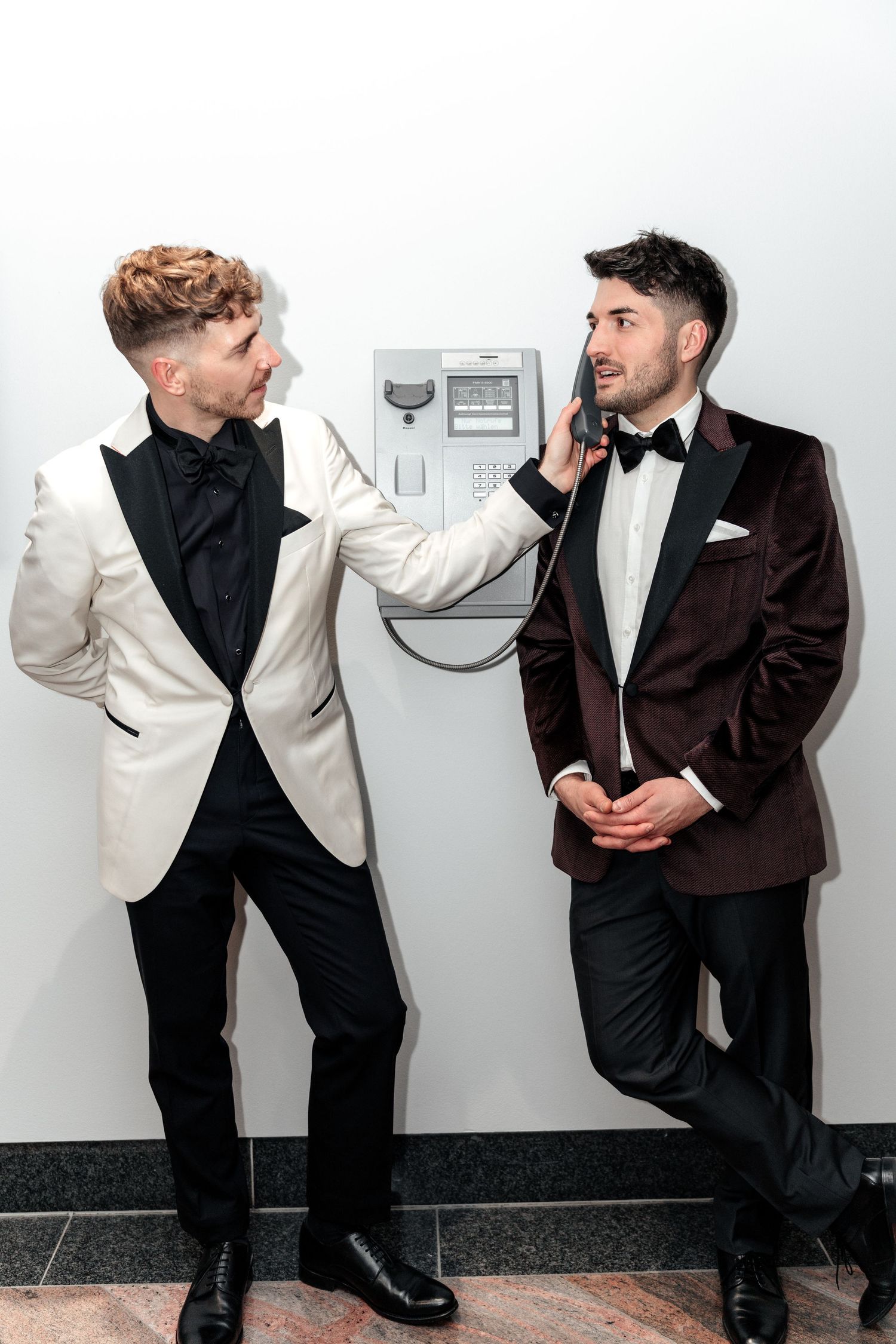 Two men in formal tuxedos pose next to each other against a wall, one wearing white and one wearing brown velvet.