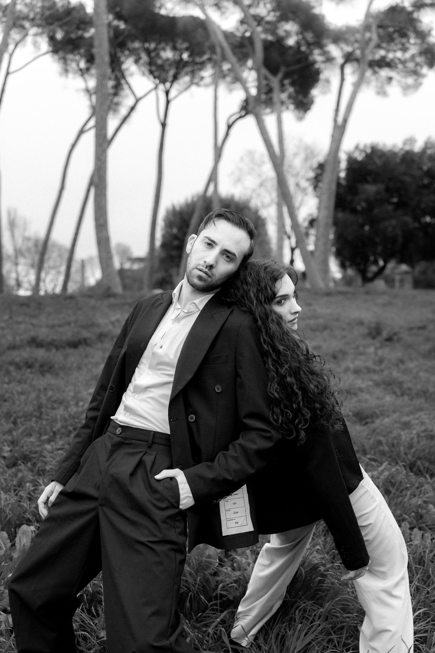 Artistic black and white portrait of a couple in contrasting outfits posing dramatically against tall trees.