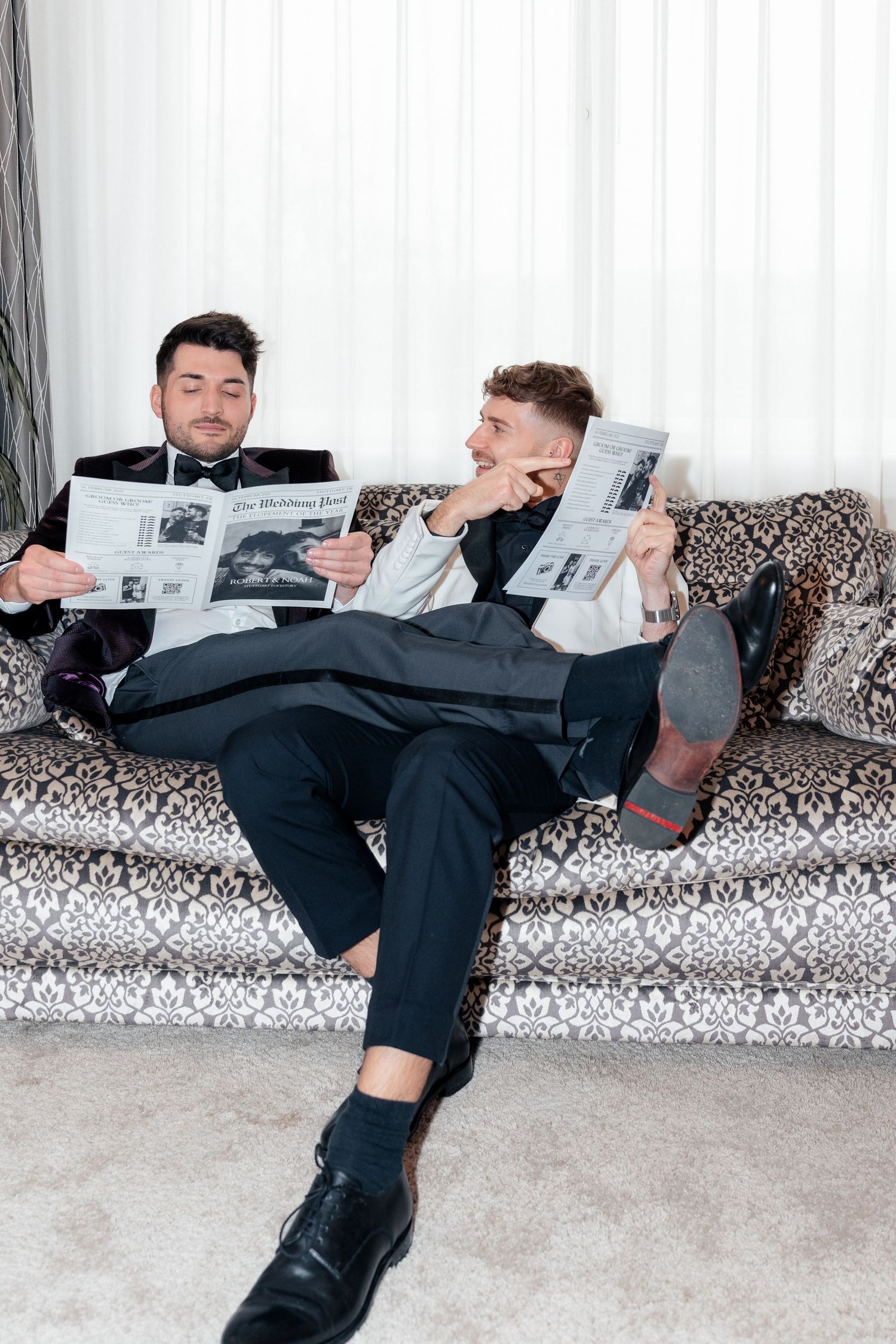 Two men in suits lounge on a patterned couch while reading newspapers in a bright, modern interior setting.