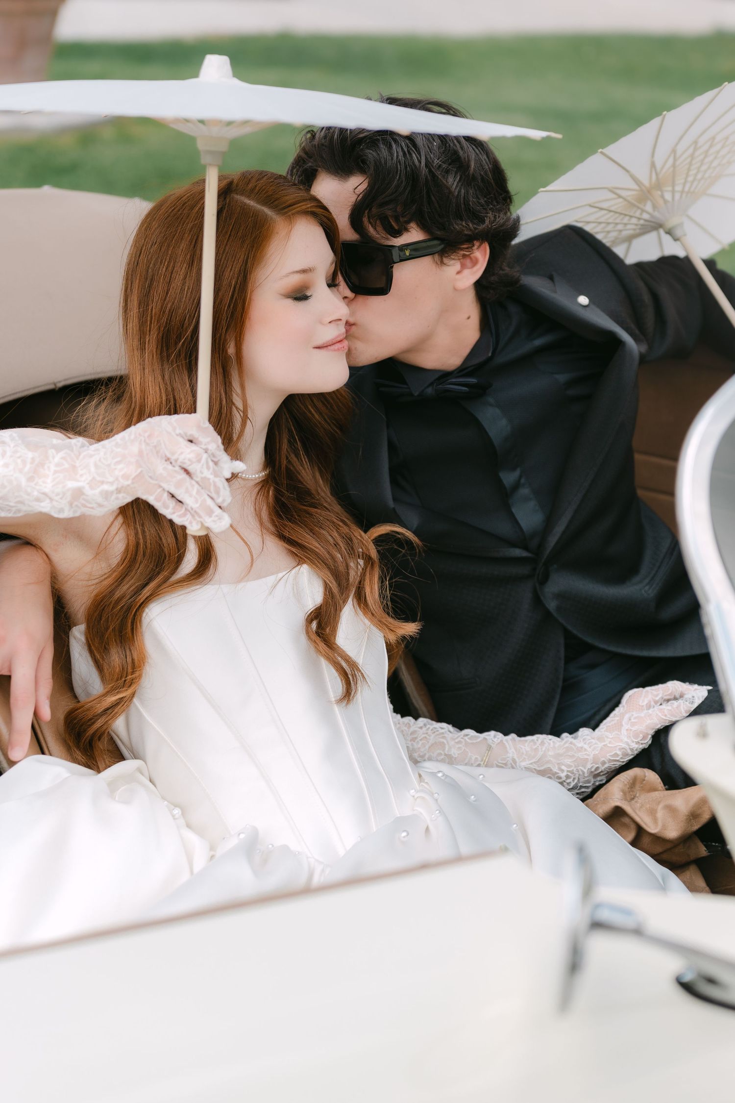 A romantic couple in wedding attire share intimate moments under a white parasol in a garden setting.