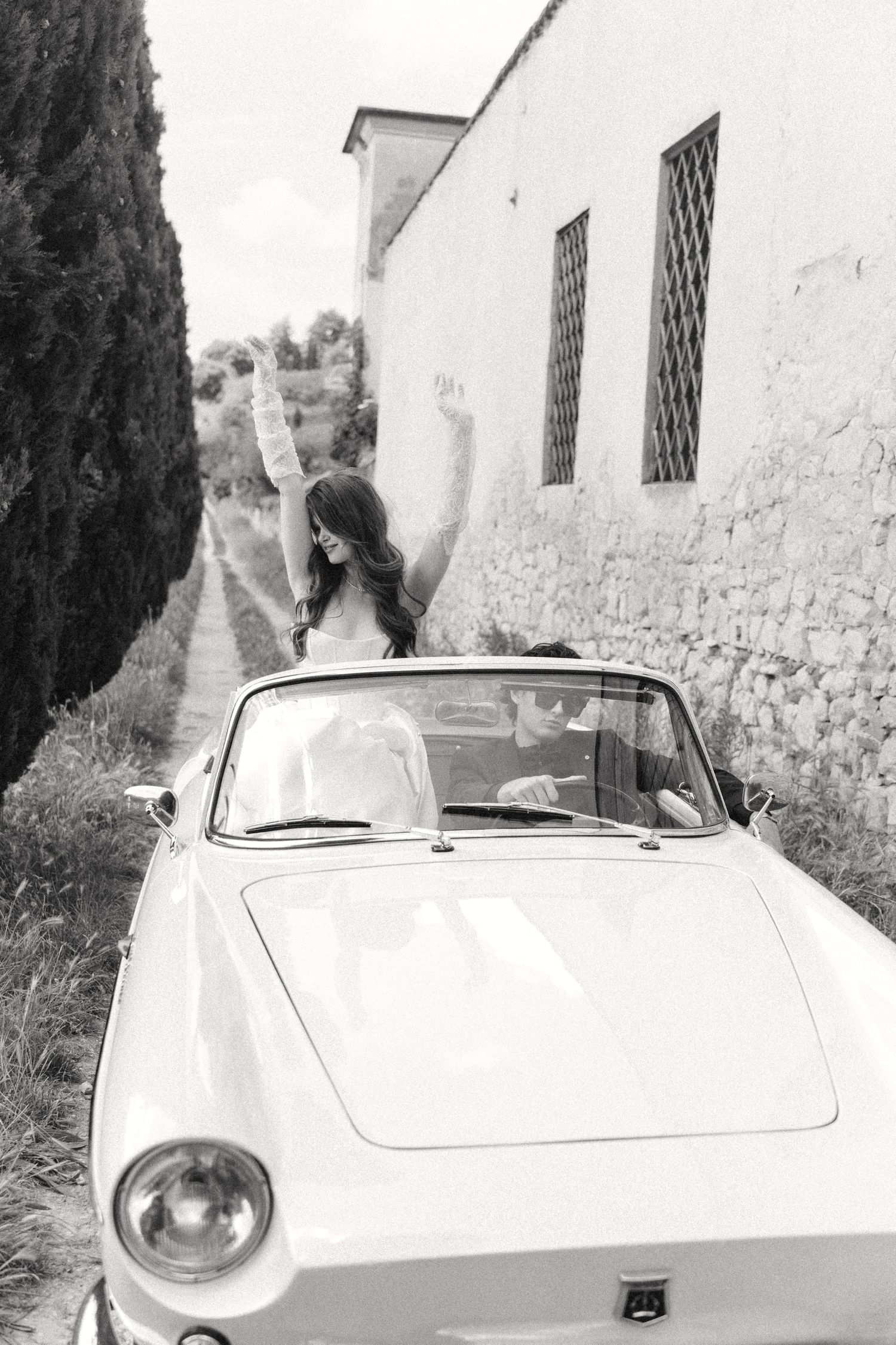 Black and white photo of someone standing in a vintage convertible car on a narrow European street with white walls.