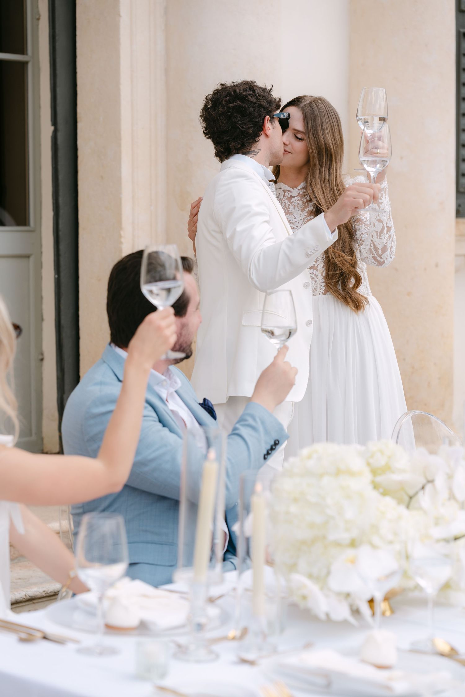 A couple shares an intimate moment at an elegant white dining table set with wine glasses and floral centerpieces. Villa Mosconi Bertani