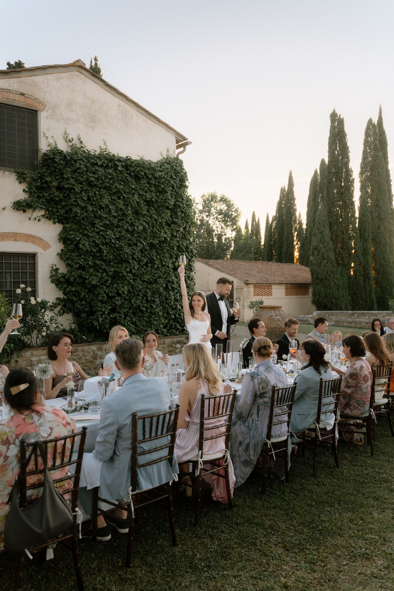 Elegant outdoor wedding reception at a Tuscan villa with guests seated at a long table, cypress trees, and ivy-covered walls.