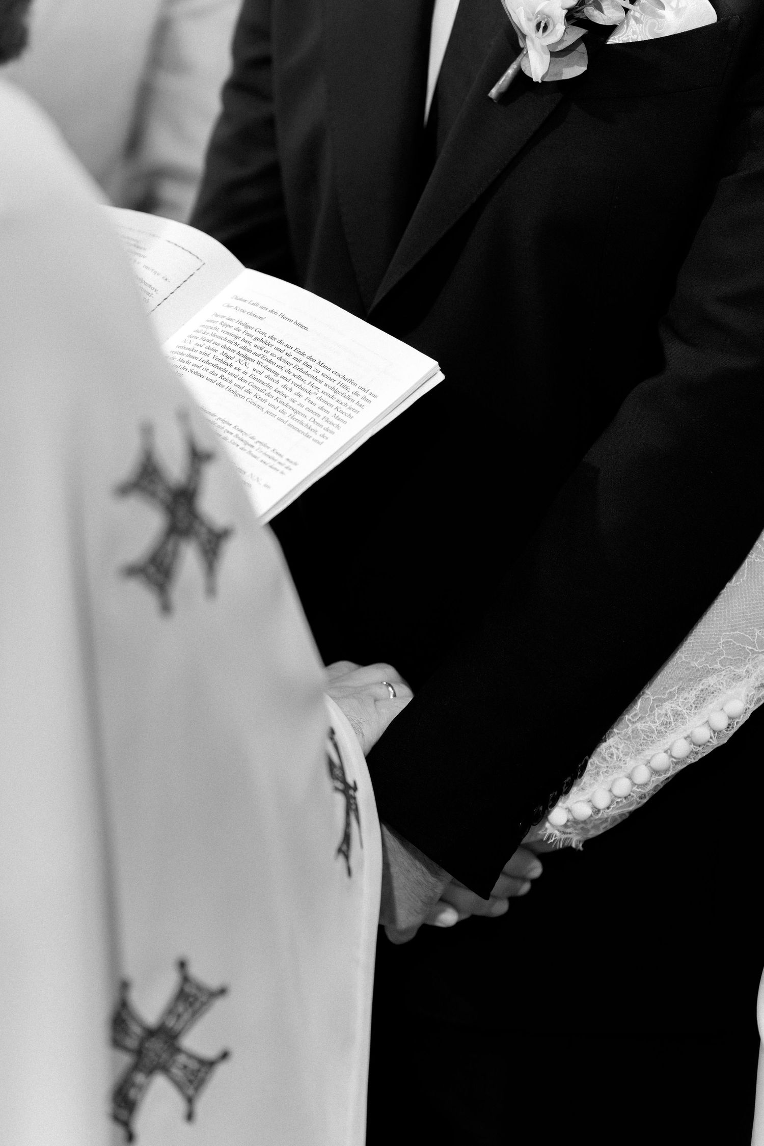 Black and white photo of couple holding hands during wedding ceremony with priest holding open Bible.