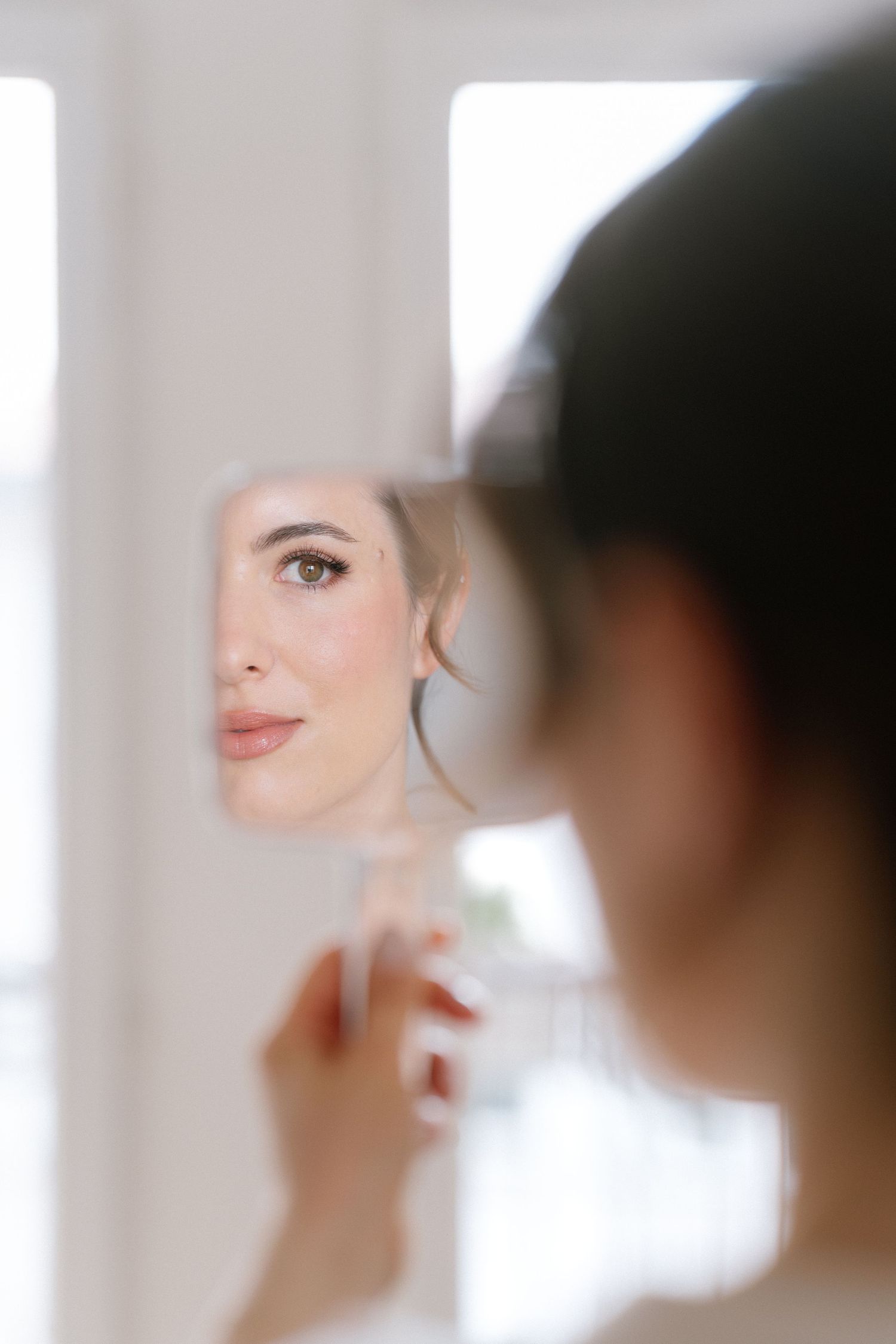 A woman holds a small mirror reflecting her face with natural makeup and elegant hair in a bright room.
