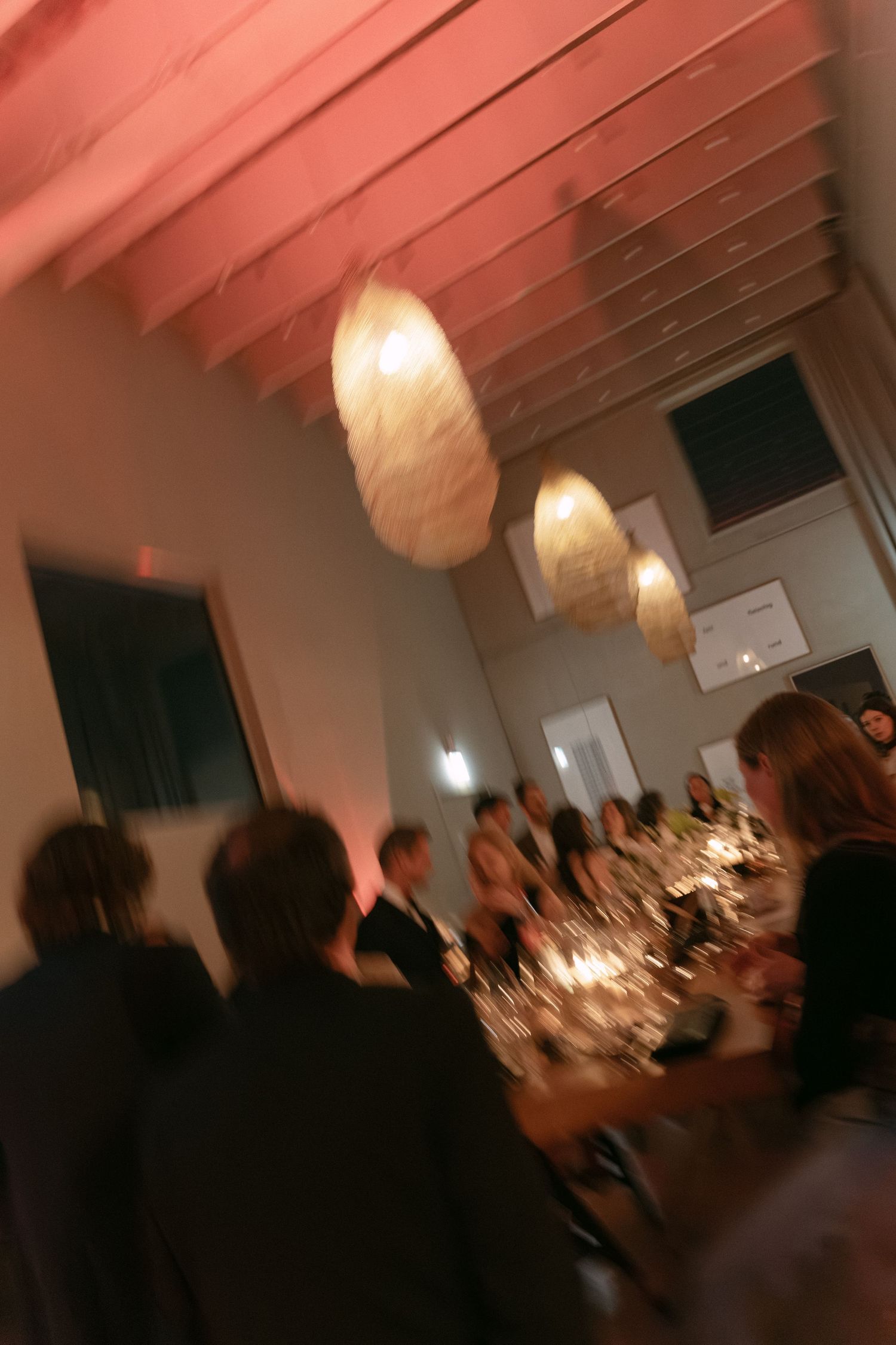Blurry candid photo of elegantly dressed guests dining at a dimly lit event venue with hanging pendant lights.