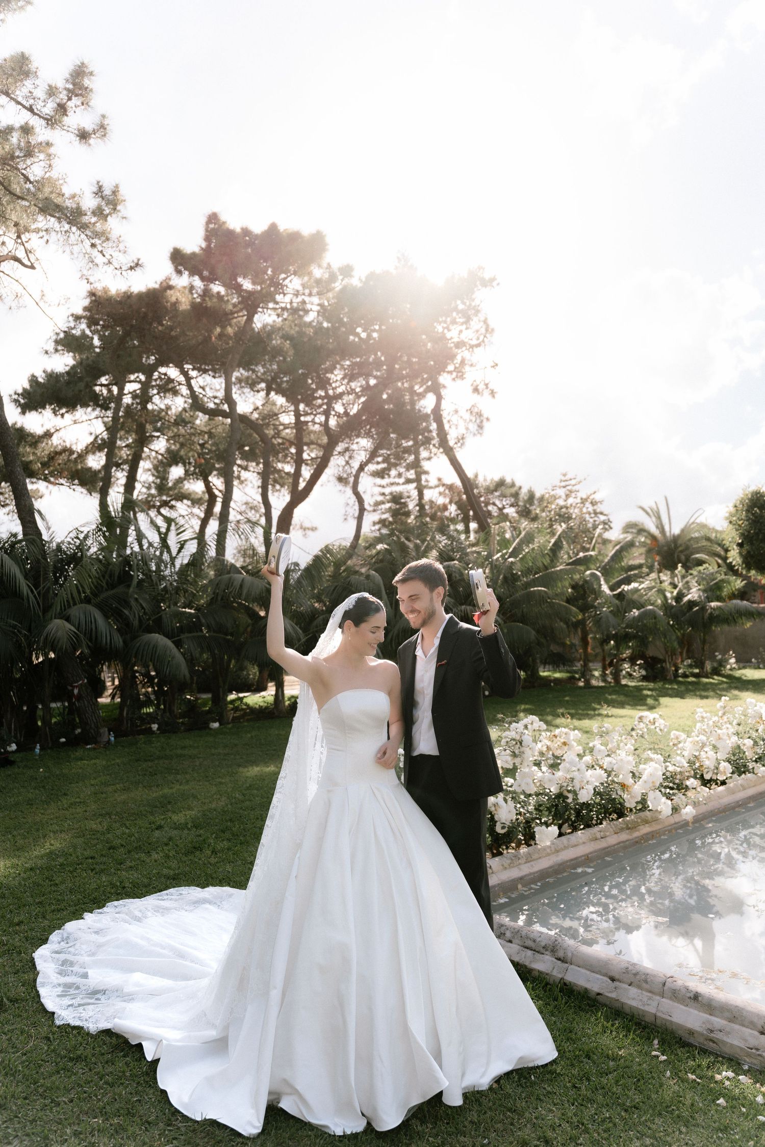 Bride in white ballgown and groom in tuxedo celebrate joyfully in a lush garden with sunlit trees.