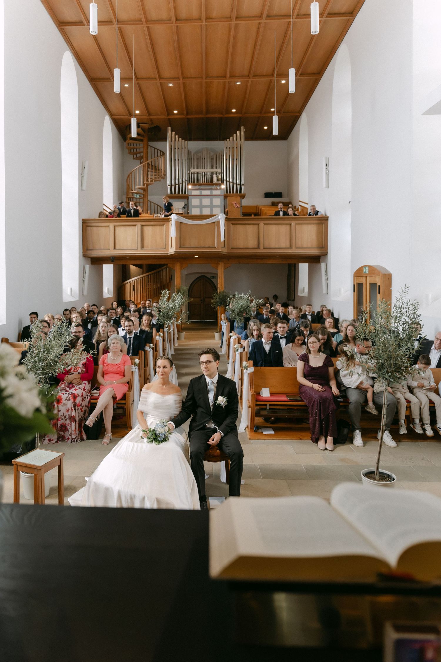 Bride and groom sit before a church altar during their wedding ceremony as guests fill the pews beneath a wooden ceiling and pipe organ.