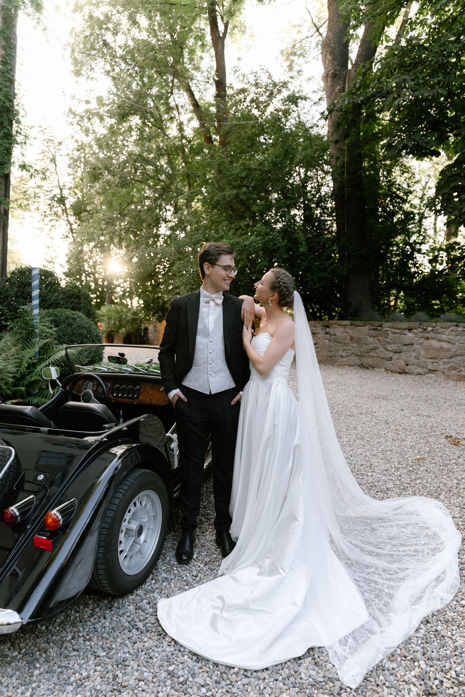 Bride in a flowing white gown with a long veil embraces a groom in a black suit beside a vintage black convertible on a gravel driveway.