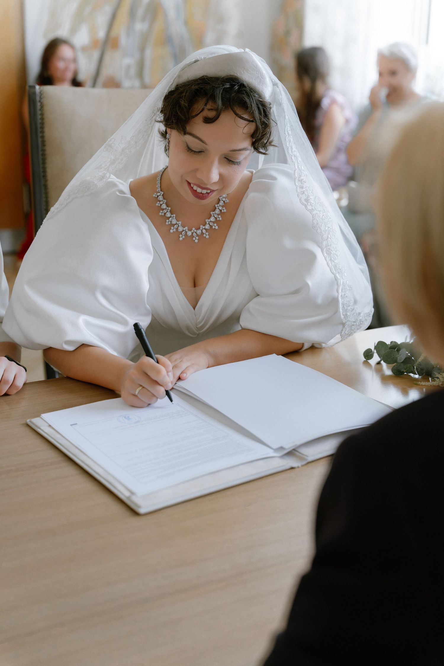 Smiling bride in white gown and veil signing marriage certificate at wooden table during wedding ceremony.