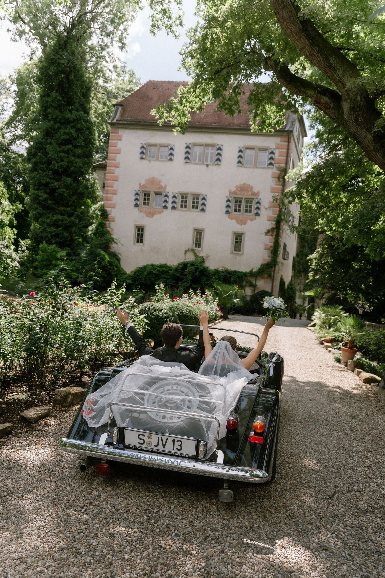 Just-married couple raise their arms in a vintage convertible with a white veil, driving down a gravel path toward a historic castle.