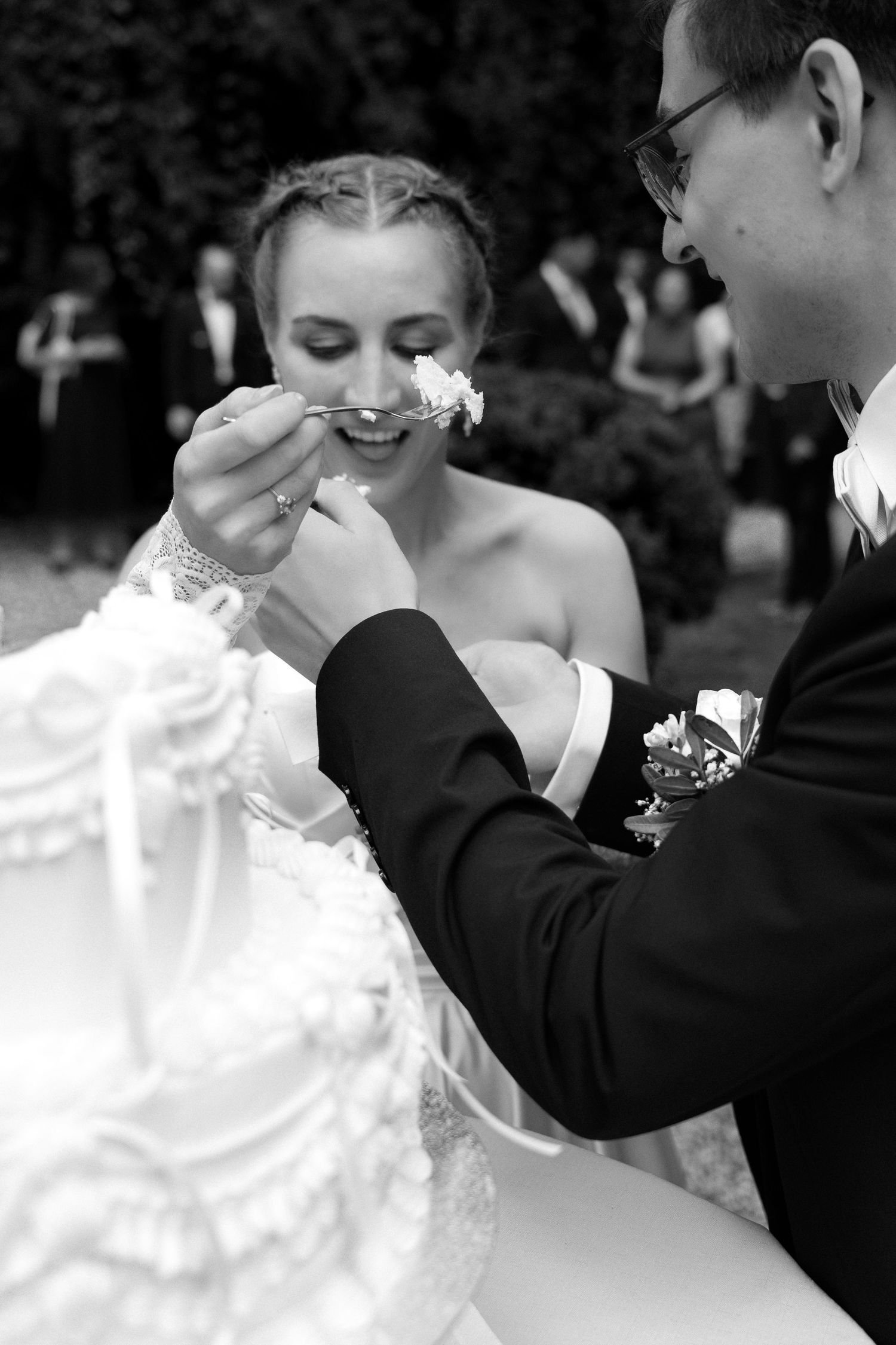 Black and white photo of a bride and groom feeding each other wedding cake at an outdoor reception with guests in the background.